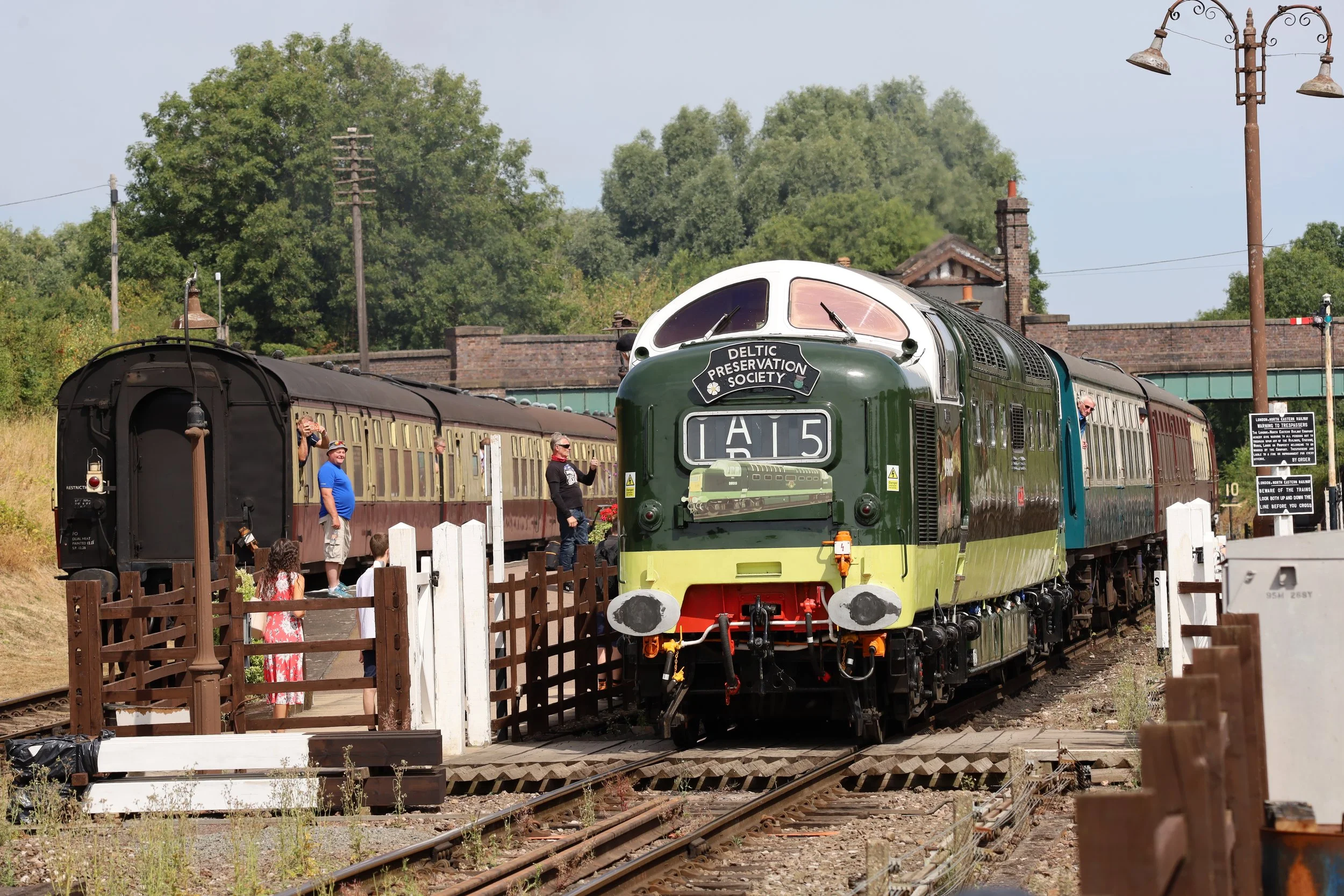 A vintage train with a green and cream locomotive and red passenger cars are on train tracks at a railway station, with people taking photos and looking at the train. There are trees, utility poles, and a bridge in the background.