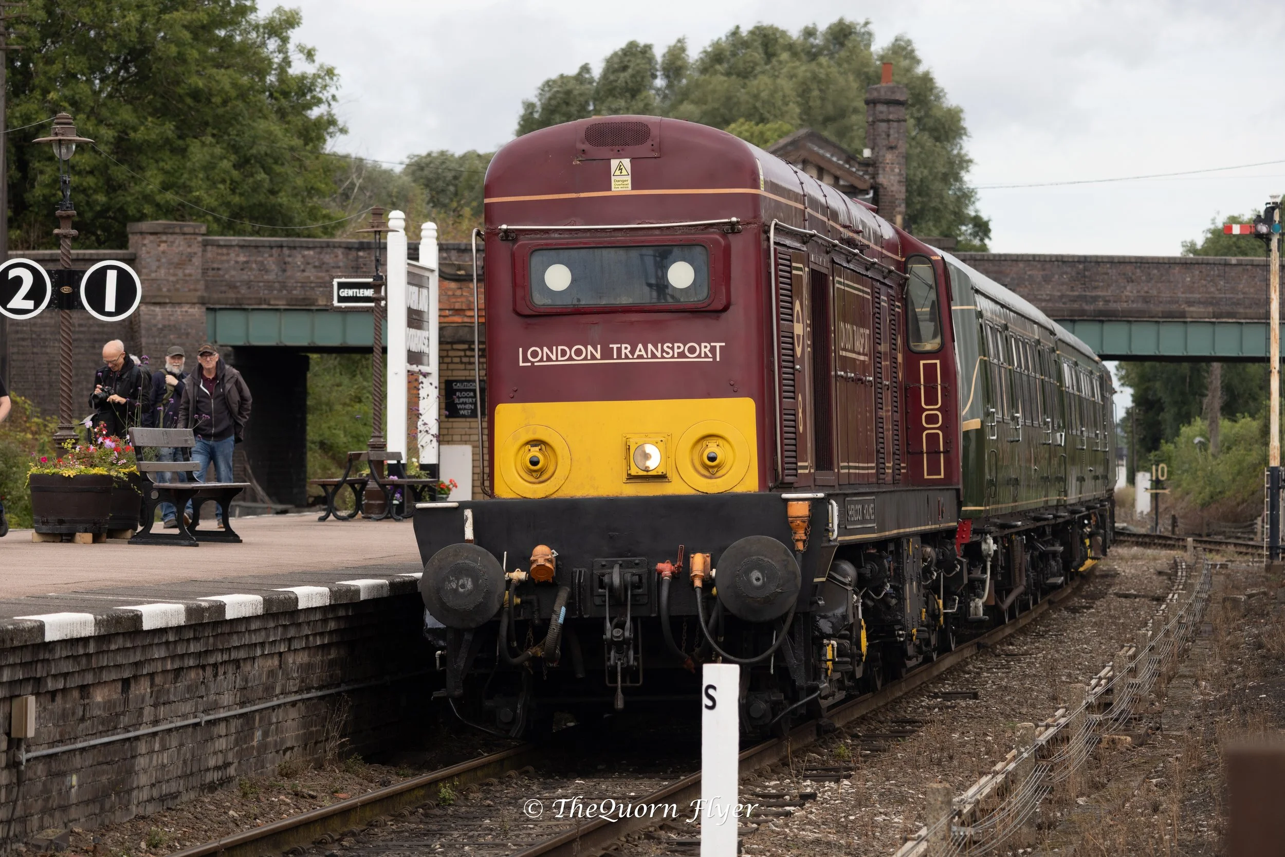 A vintage London Transport train at a station with a sign indicating platform numbers 1 and 2. There are people waiting on the platform, and the station is built with brick walls and has a bridge crossing over the tracks.