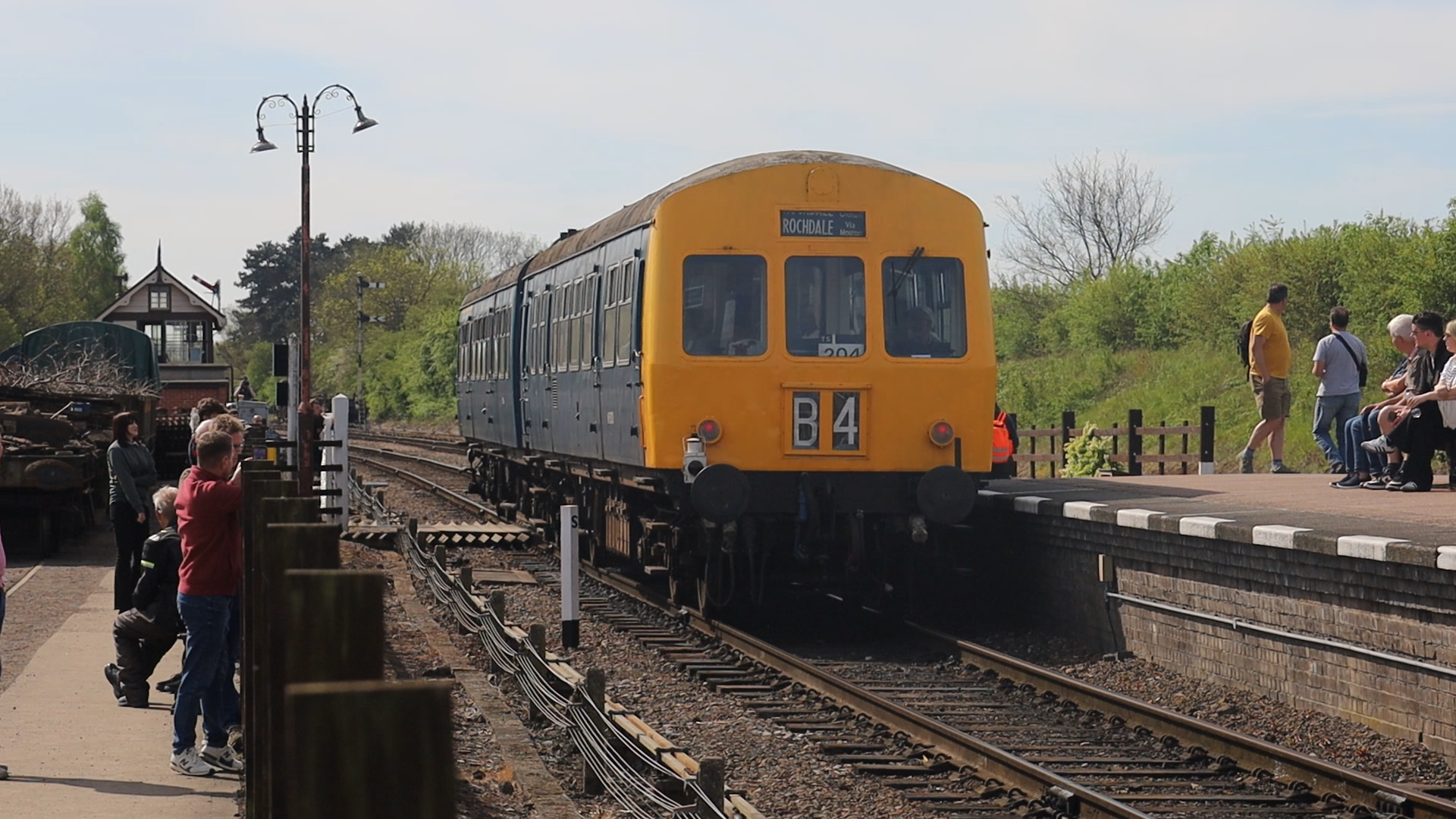 People standing and sitting on a train platform watching a yellow and blue train arrive. The train displays 'Rochdale' on its destination sign, surrounded by trees and a small station building.
