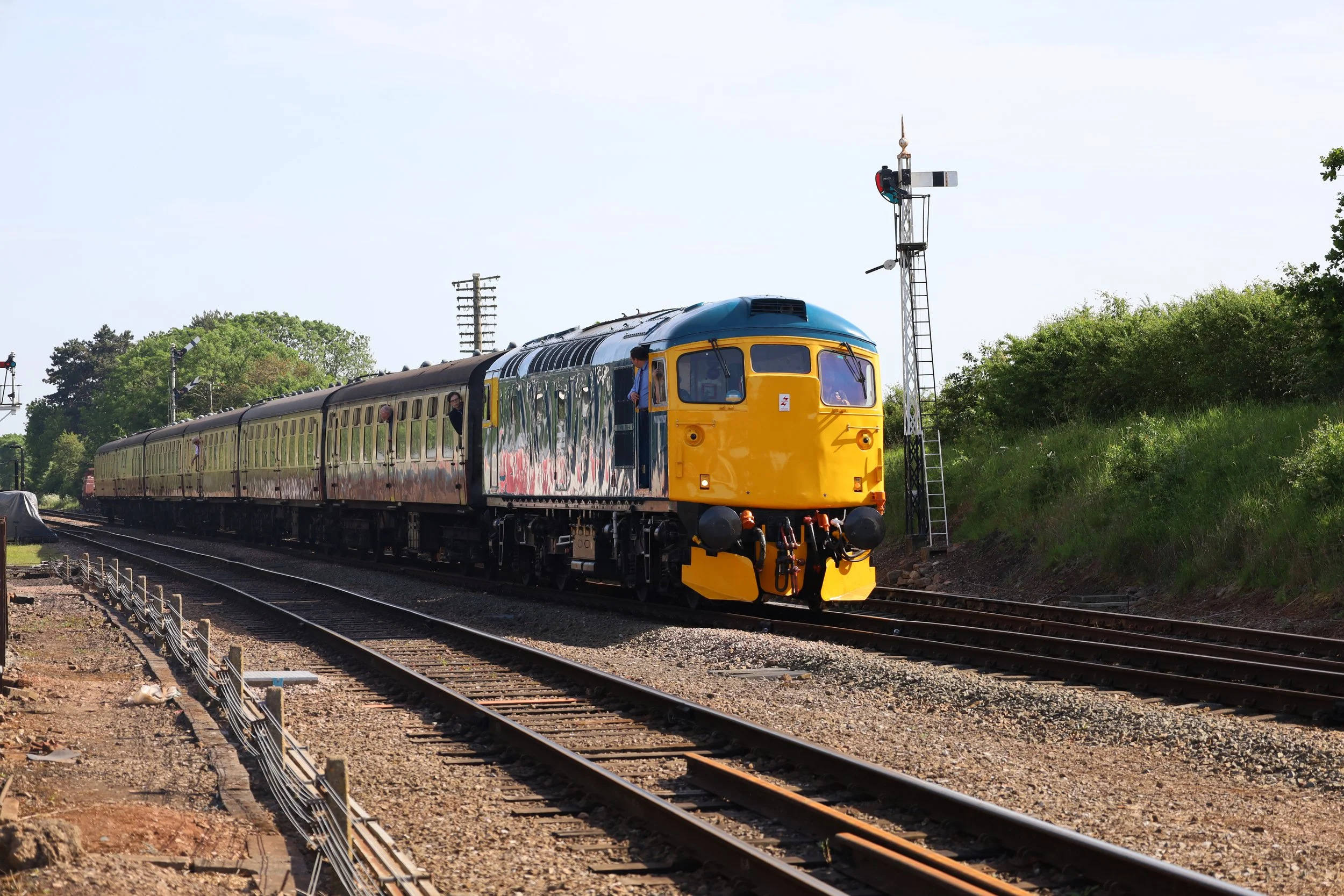 A yellow and blue train moving along railway tracks with a railway signal tower nearby, surrounded by green trees under a clear blue sky.