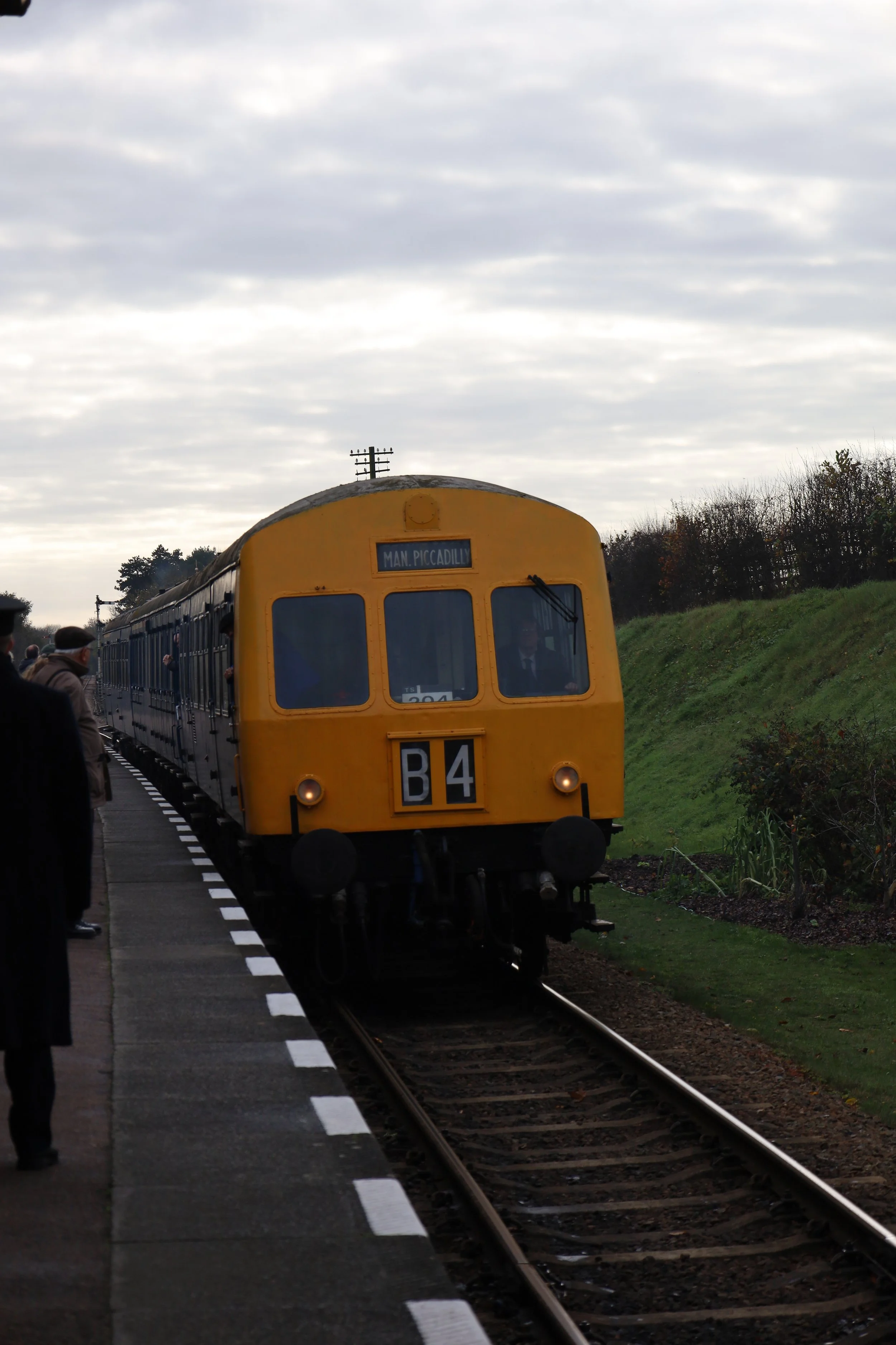 A vintage yellow train at a station platform with a sign indicating 'MAN. PICCADILLY'. Several people are waiting on the platform side.
