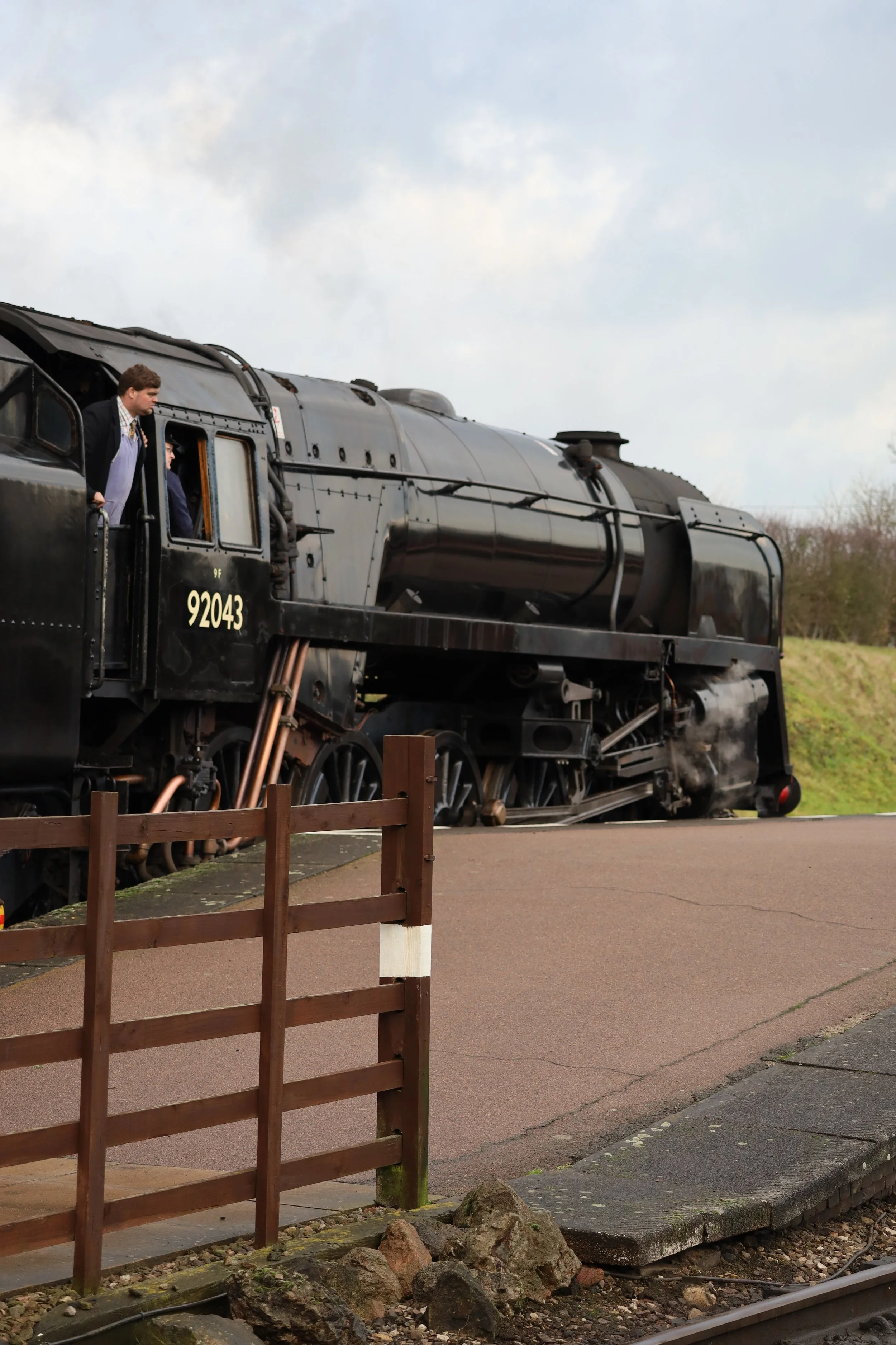 A vintage black steam locomotive train with the number 92043 on its side, seen outdoors on a cloudy day, with a man dressed in old-fashioned attire leaning out of the window.