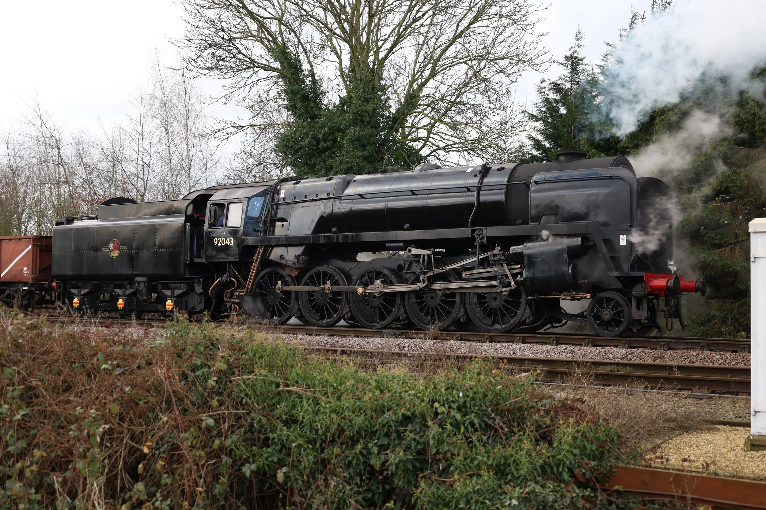 A black steam locomotive with the number 92043 on the side, emitting white steam, on train tracks in a rural outdoor area with trees in the background.