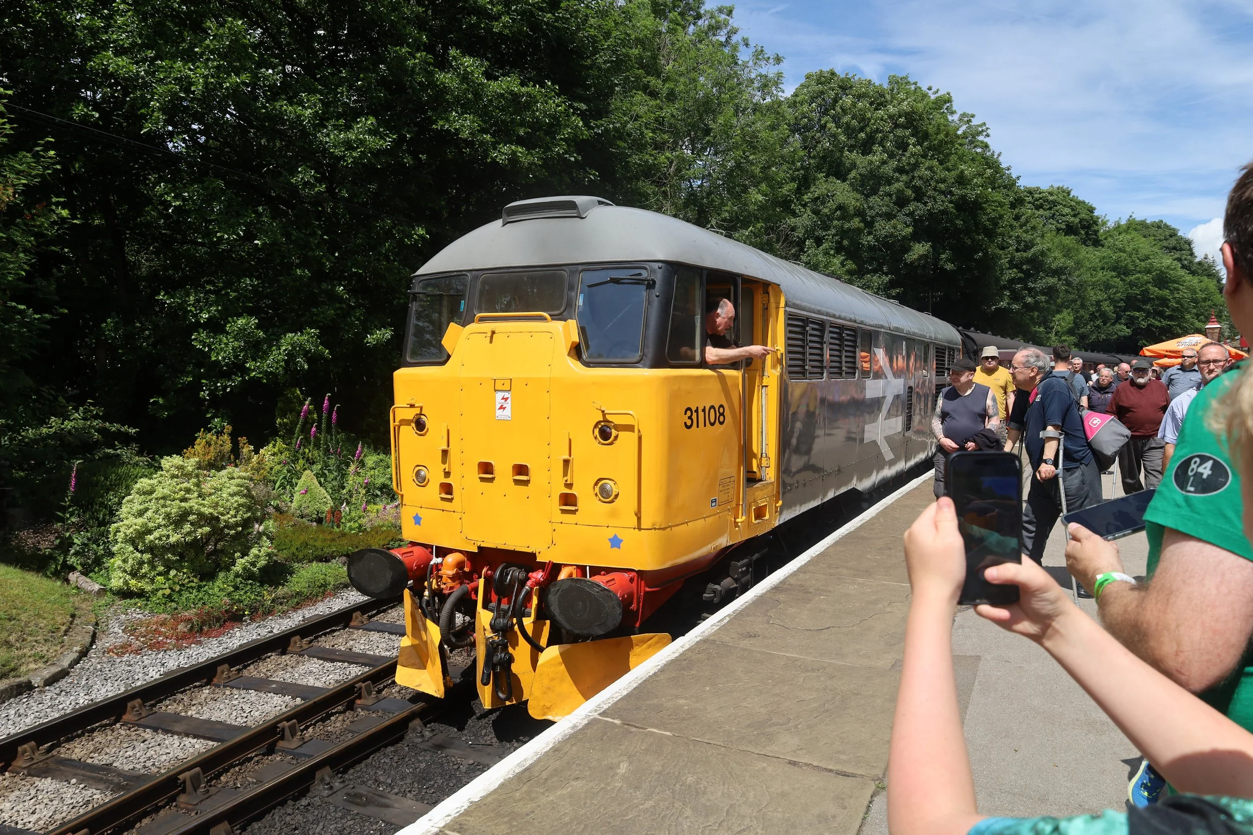 A restored vintage train engine with the number 31108 on the front, yellow and black in color, is on a track with a group of people gathered around it, taking photos. The scene is outdoors with lush green trees on the side and a partly cloudy sky.