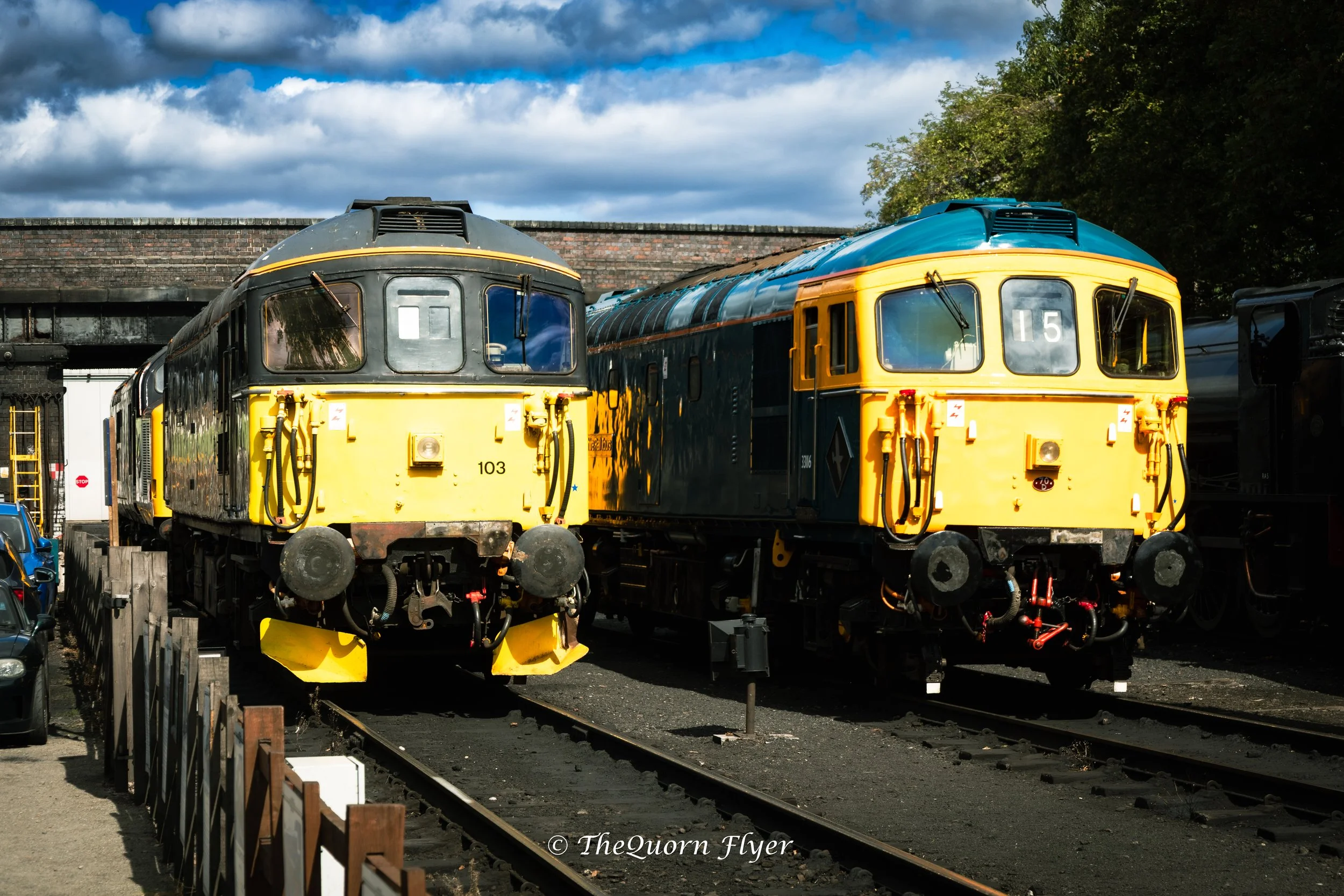 Two vintage train locomotives, one black and one yellow, parked on train tracks outdoors beneath a partly cloudy sky with green trees to the side.