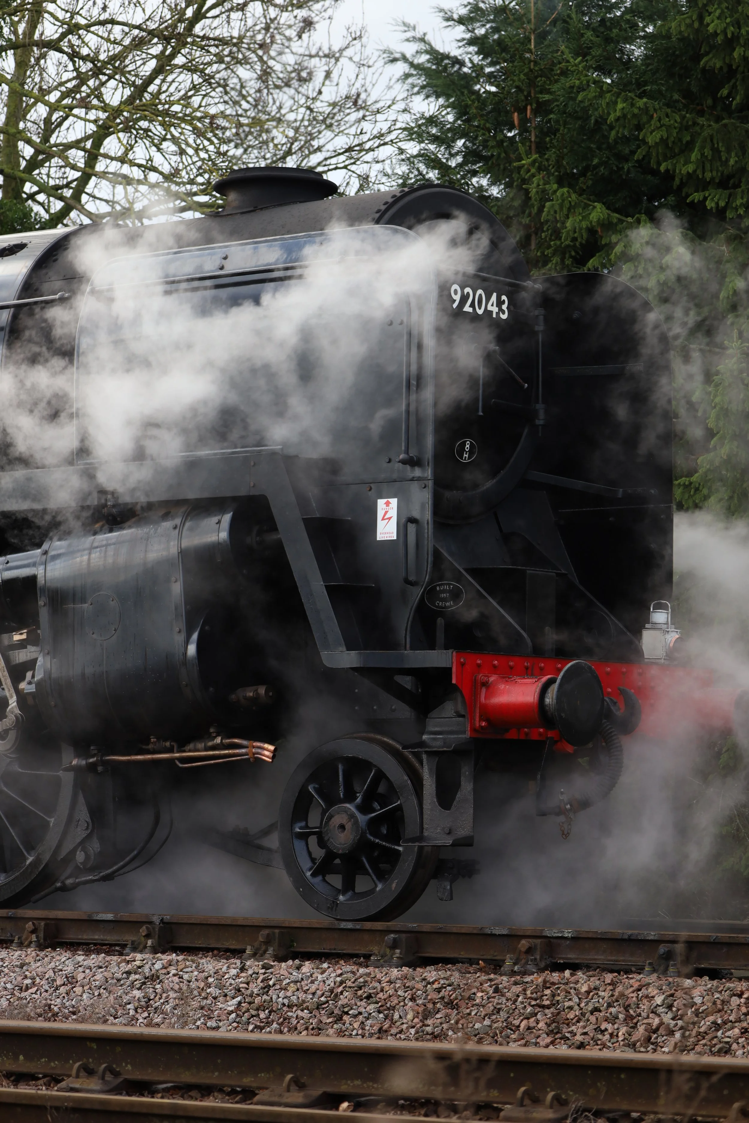 A vintage black steam locomotive emitting smoke and steam, with the number 92043 on the front, traveling on railway tracks surrounded by trees.