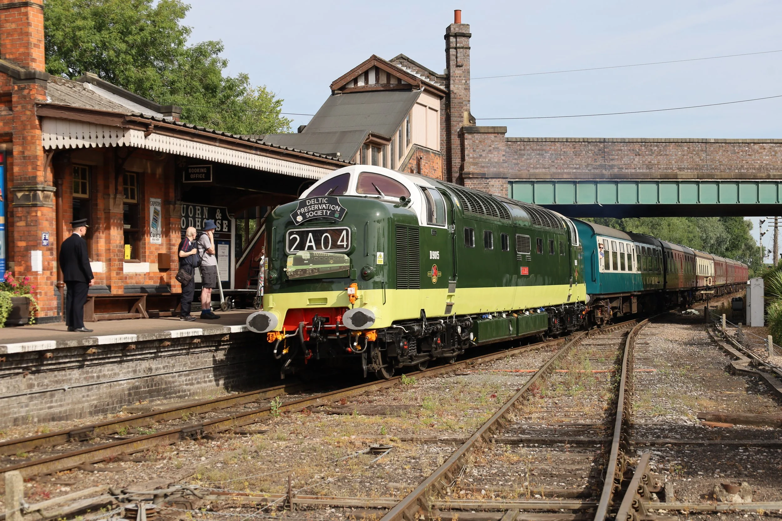 A vintage train is stopped at a station platform with people awaiting departure. The station building has a sign that reads 'Booking Office' and a poster advertising 'Capslock & House.' The train has a green and cream color scheme with the sign 'Deltic Preservation Society' on the front. The scene is outdoor with trees and a blue sky in the background.