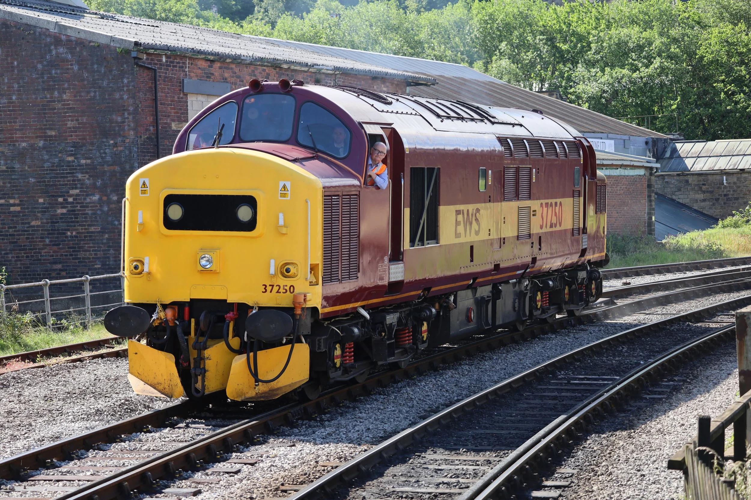 A vintage industrial locomotive painted in yellow and maroon, moving along railway tracks with trees and a brick building in the background, featuring two people visible in the cab