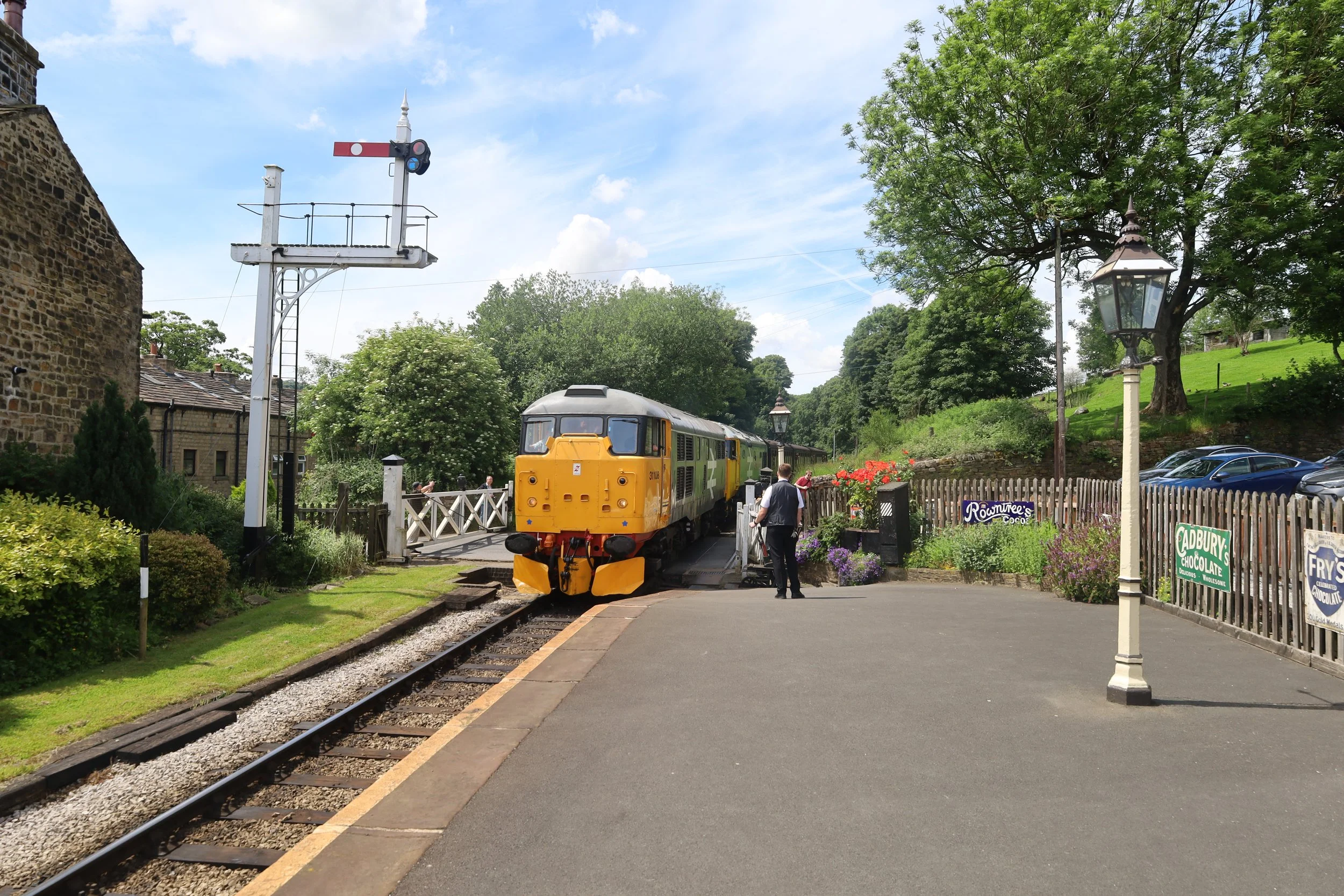 A yellow train is stopped on the tracks at a small, scenic station with lush green trees and buildings around. A person is standing near the train, and a signal light is visible on a tall pole. There are parking spaces with cars and signs for local businesses.