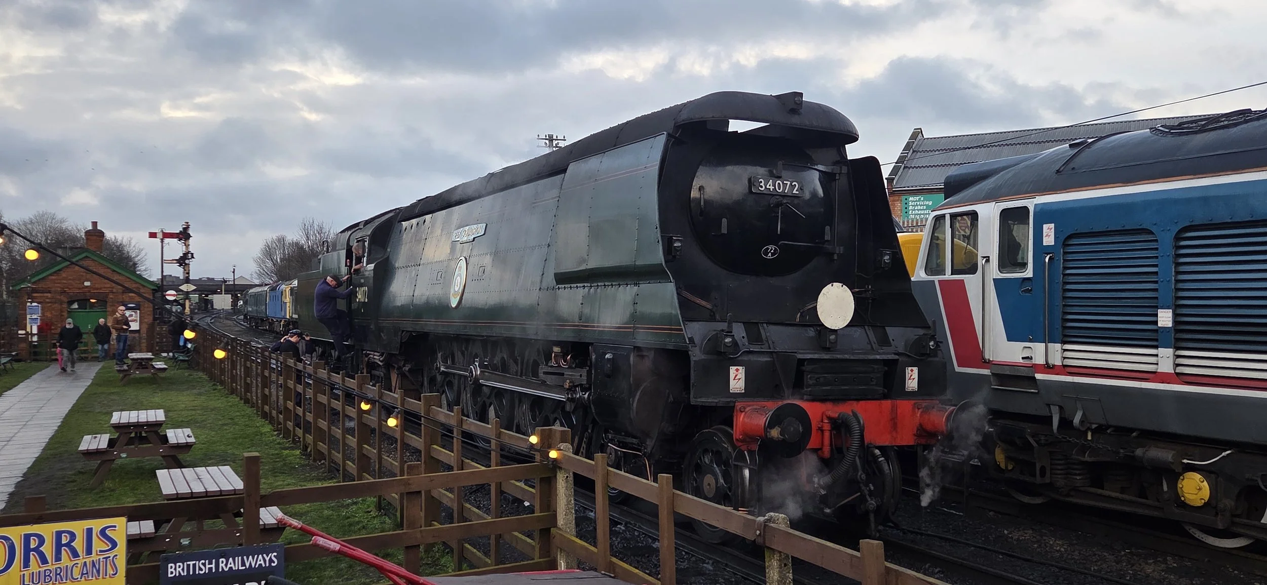 A vintage British steam locomotive on a track with a few people around, some taking pictures. The scene is outdoors, with a cloudy sky and small wooden picnic tables nearby.