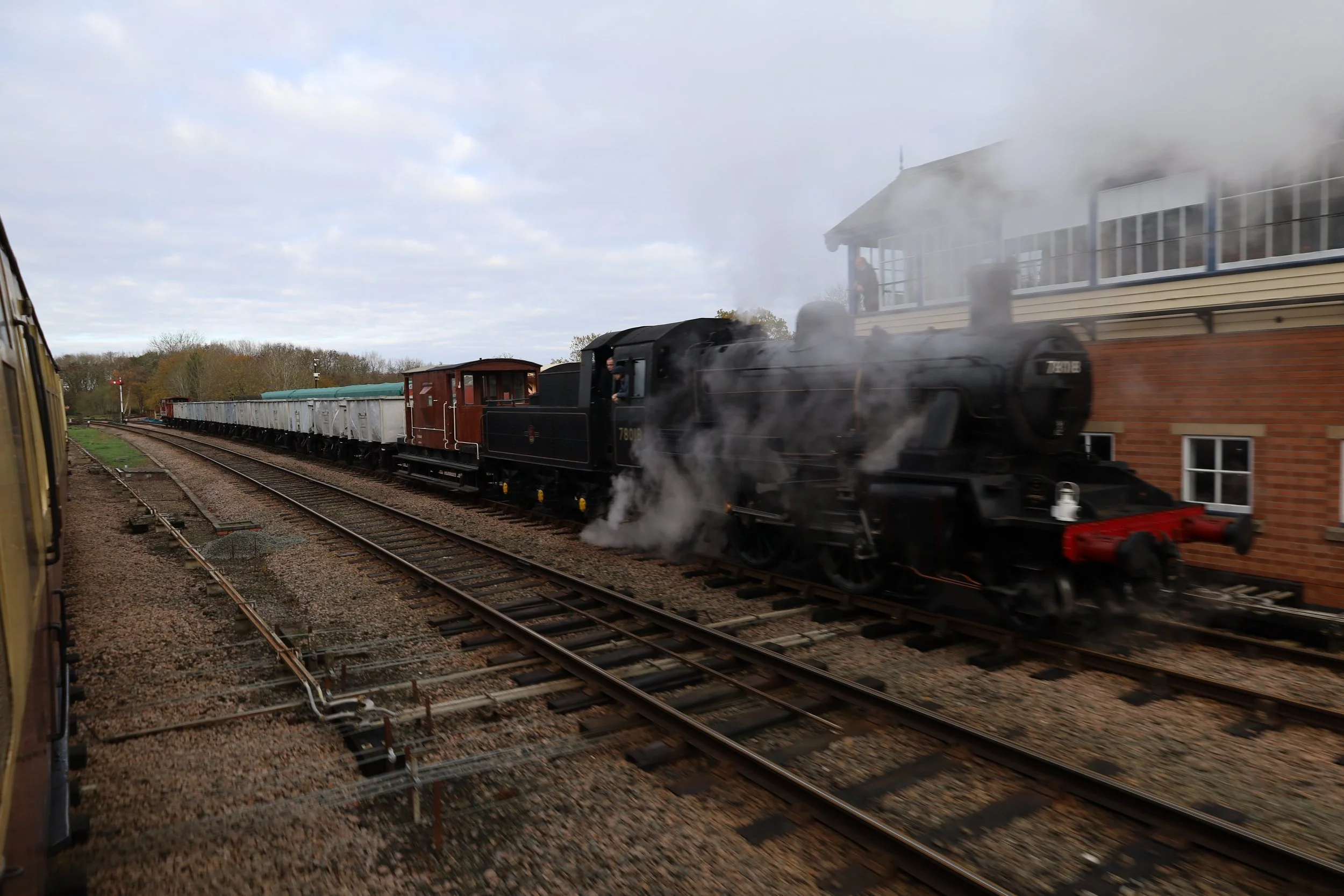 A black steam train moving on railway tracks, with smoke billowing from its chimney, passing by a brick building and a station platform with a person standing on it, in a rural setting with trees in the background.
