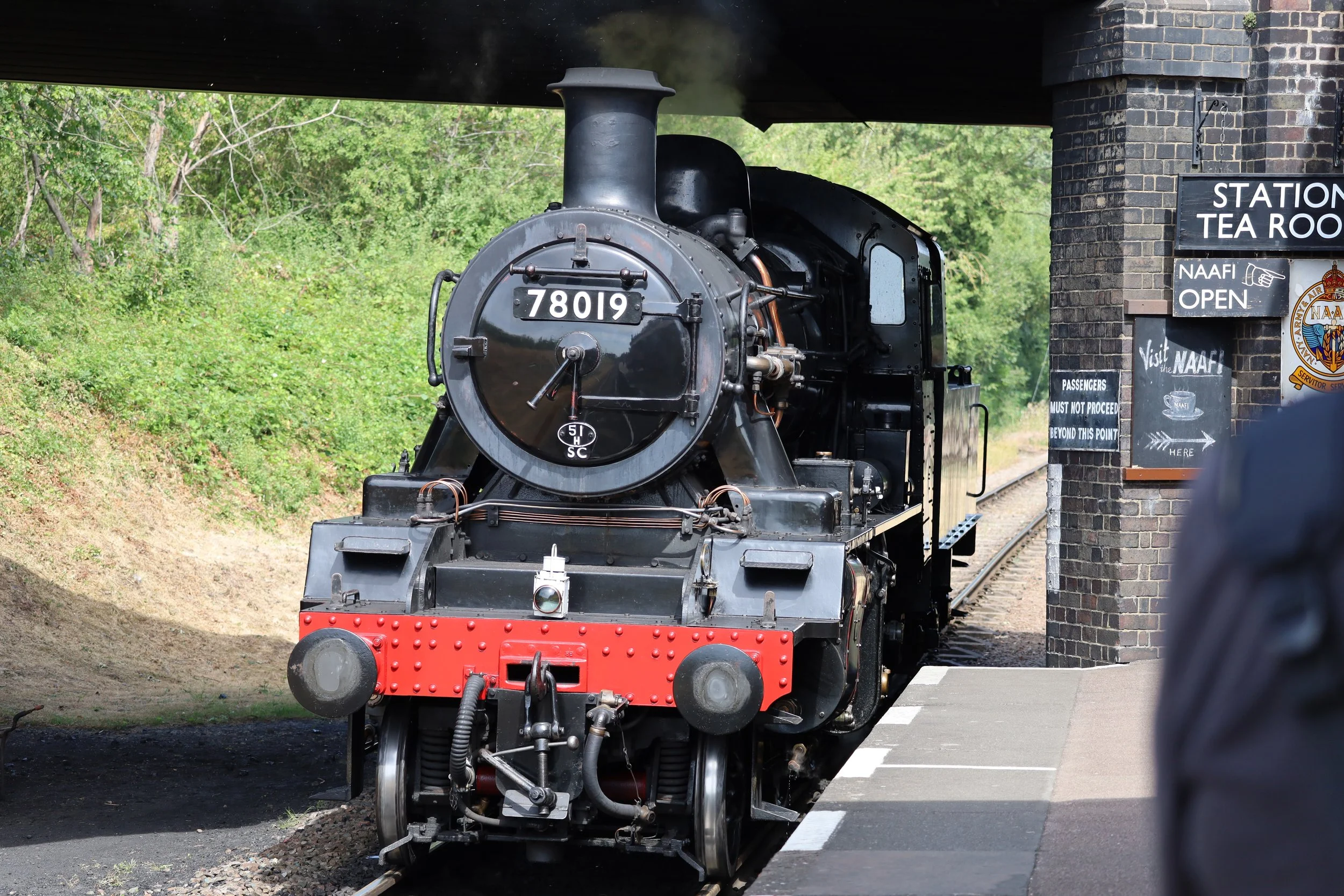 A vintage black steam locomotive with the number 78019 on the front, emerging from under a bridge near a brick station building with a sign that reads 'Station Tea Room.' Green trees and foliage are visible in the background.
