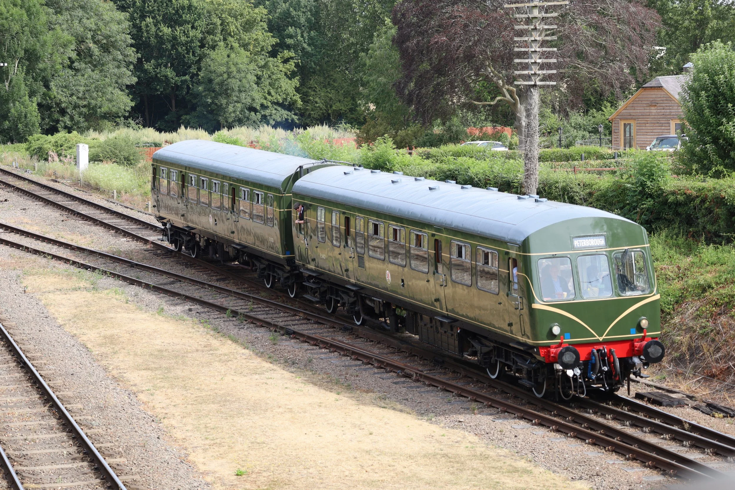 A vintage green train on railway tracks surrounded by lush green trees and a small wooden building in the background.