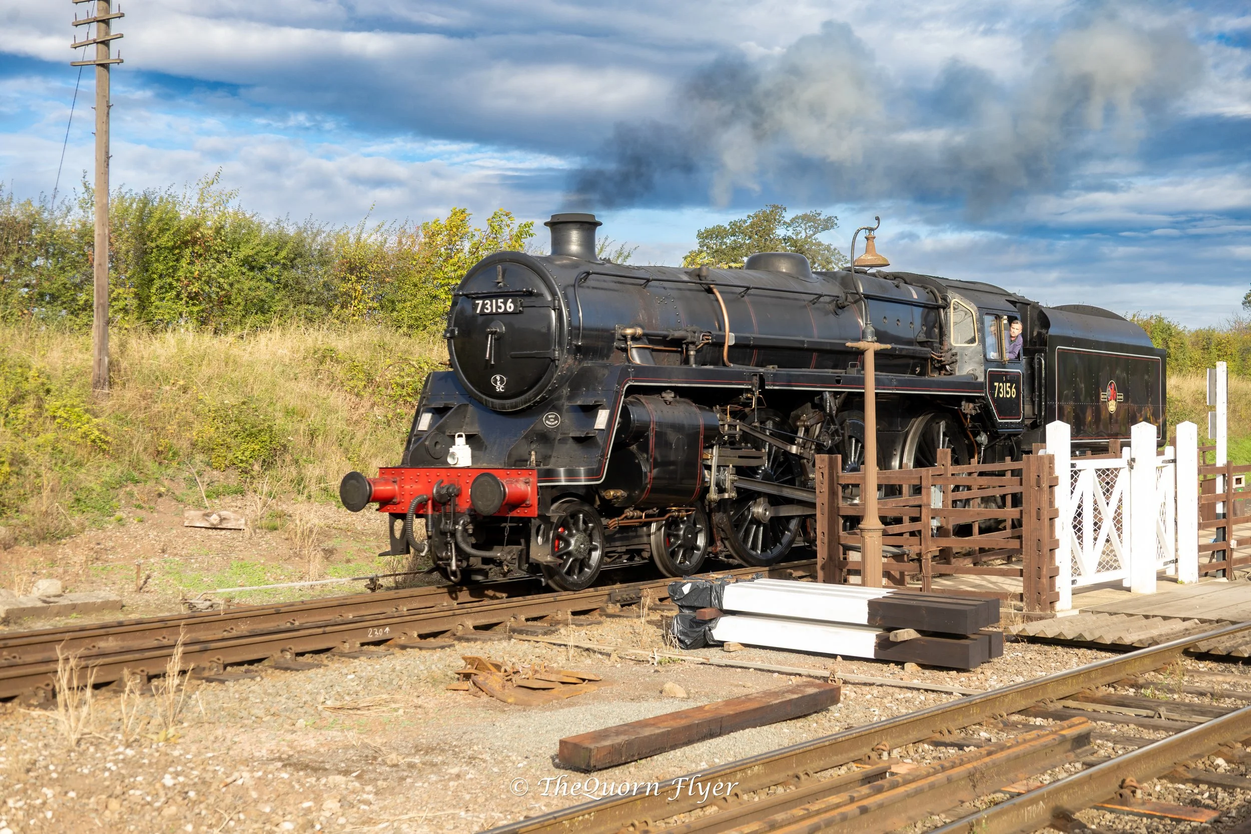 A vintage black steam locomotive with the number 73156 on the front and sides, positioned on train tracks with some greenery and trees in the background, under a partly cloudy sky.