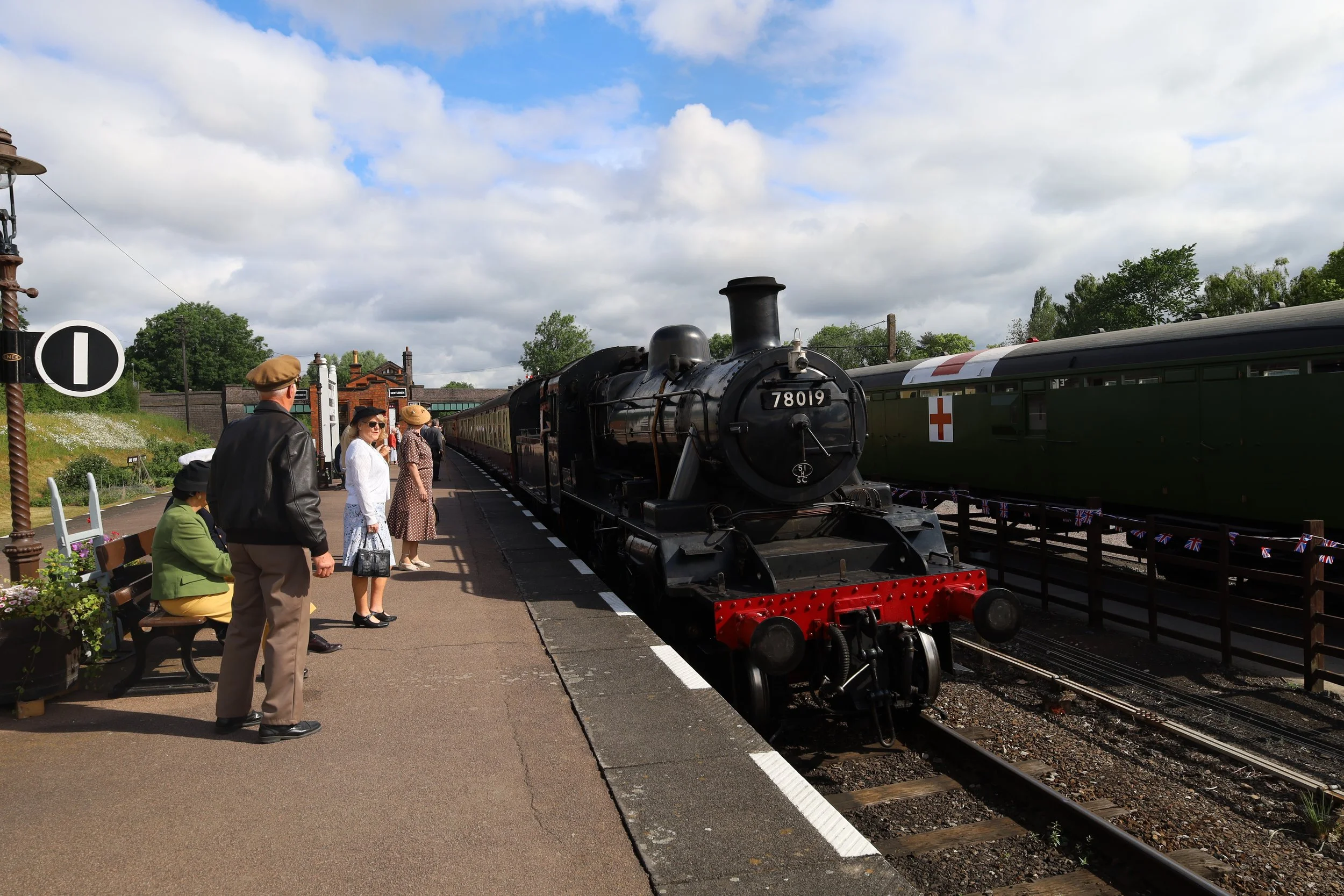 People standing and sitting on benches near a black steam locomotive train with the number 78019, at an outdoor train station on a partly cloudy day.