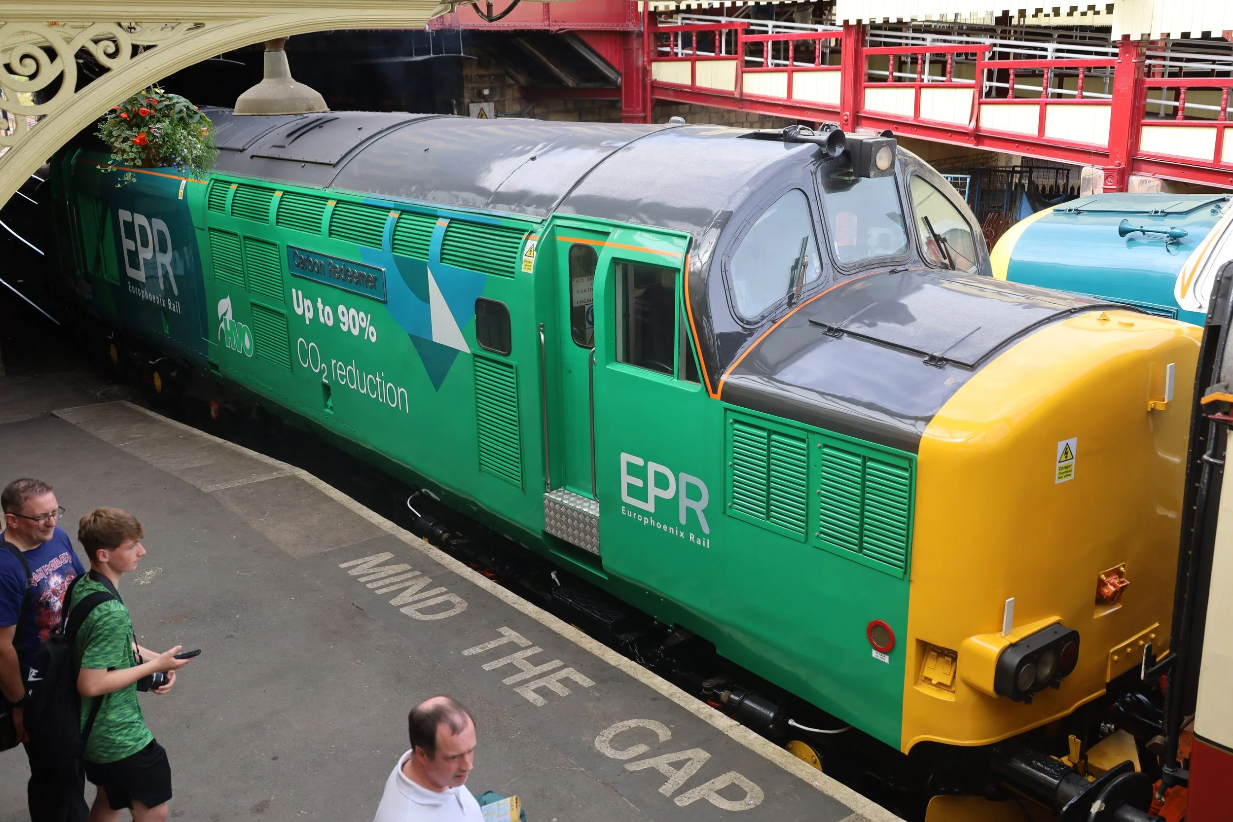A green and yellow train with the logo EPR on its side parked inside a station. Several people are standing nearby, some looking at the train and others using their phones.