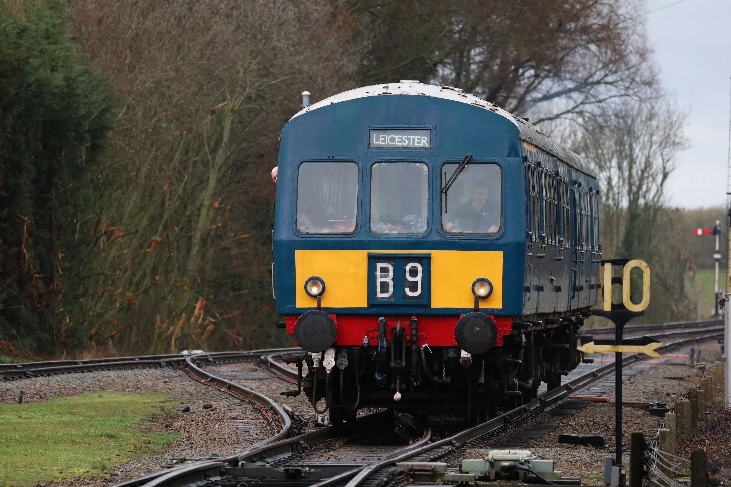 A vintage blue train engine labeled 'LEICESTER' traveling on railway tracks through a rural area with trees and cloudy sky in the background.