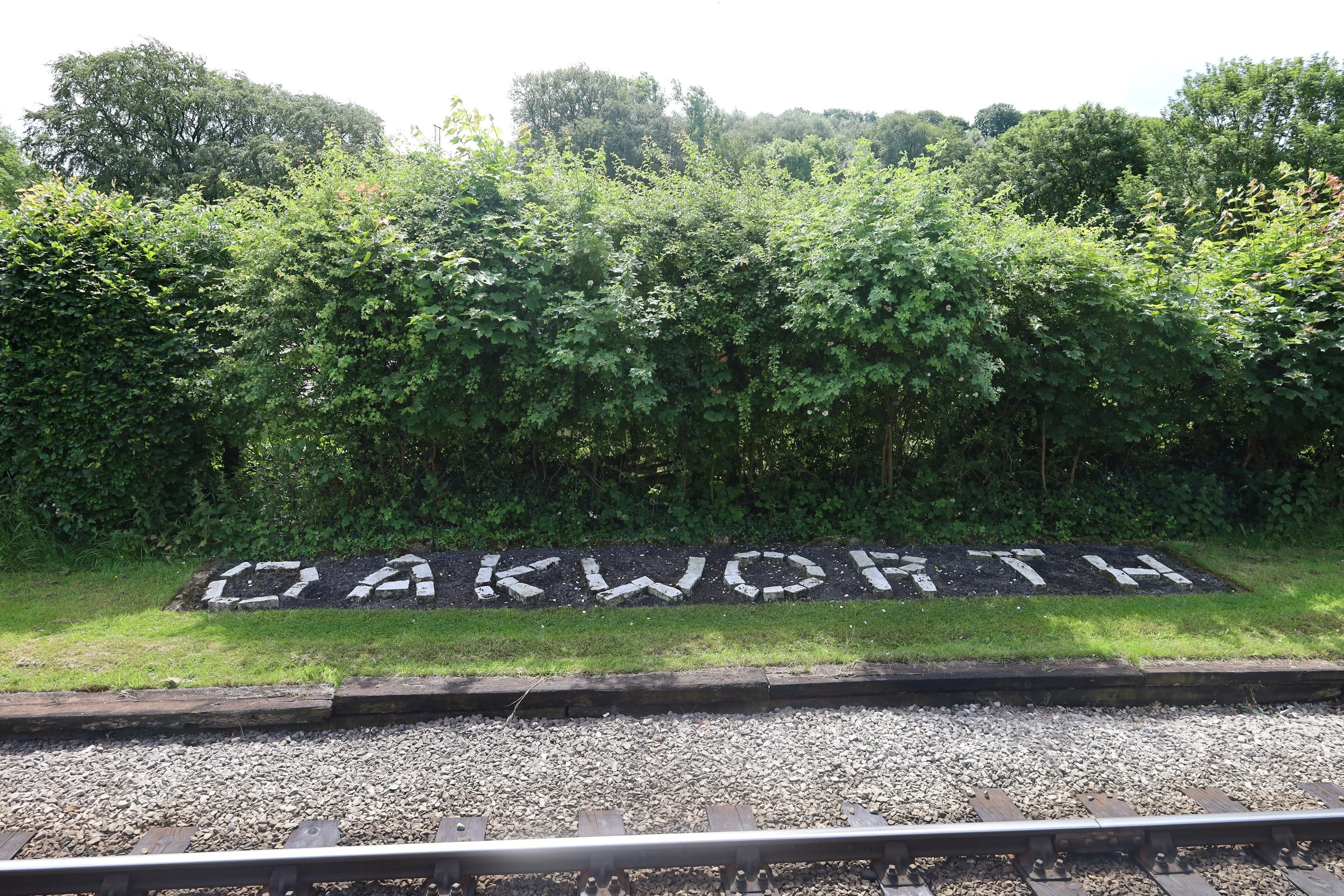 View of green bushes and trees with a railway track in the foreground and the word 'ACKNOWLEDGE' spelled out in white stones on dark soil.