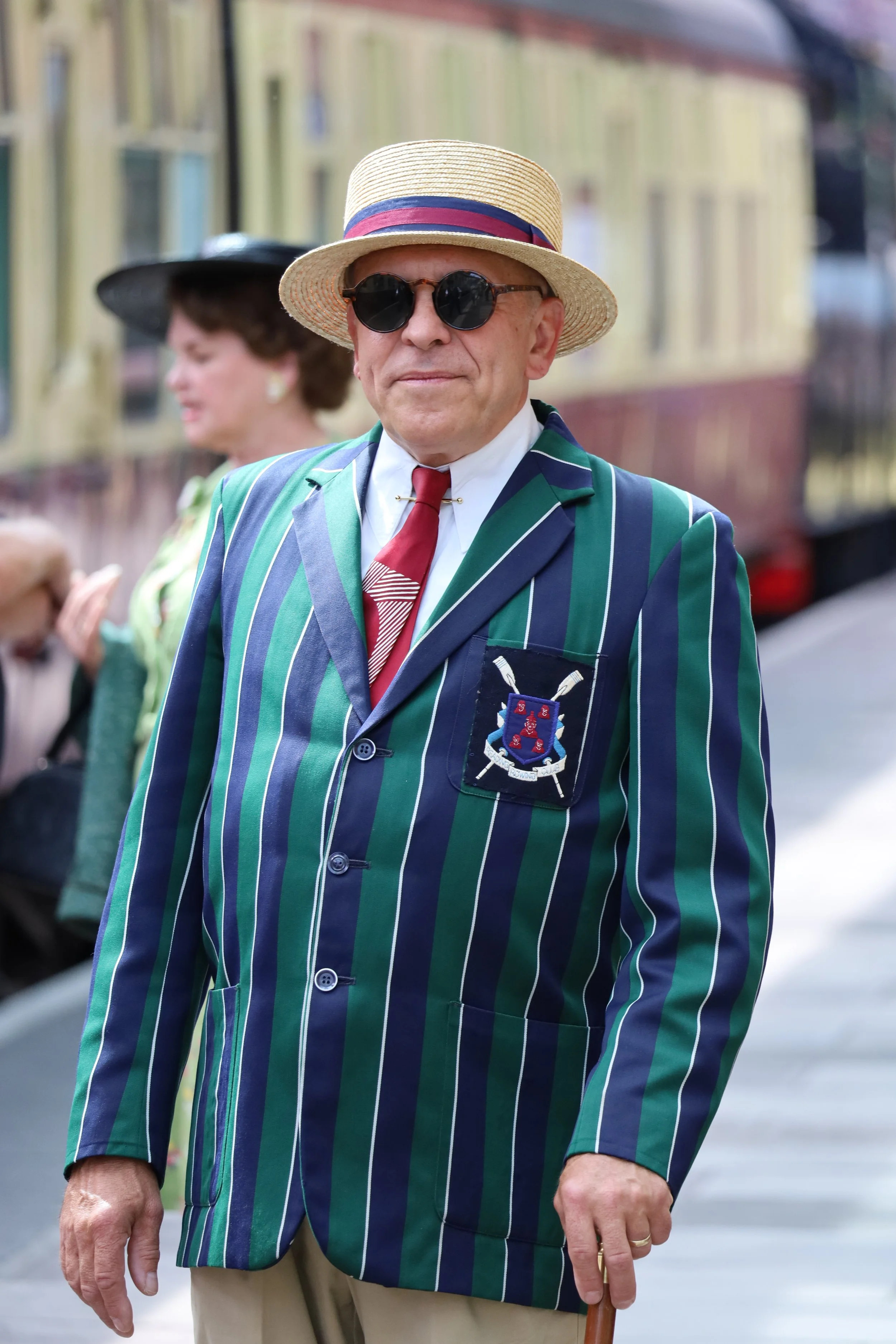 A man wearing a straw hat with a red, white, and blue band, black round sunglasses, a striped green, blue, and navy blazer with a crest on the pocket, a white shirt, a red and white striped tie, and khaki pants, standing outdoors near a yellow train with a woman in a black hat and a green outfit in the background.