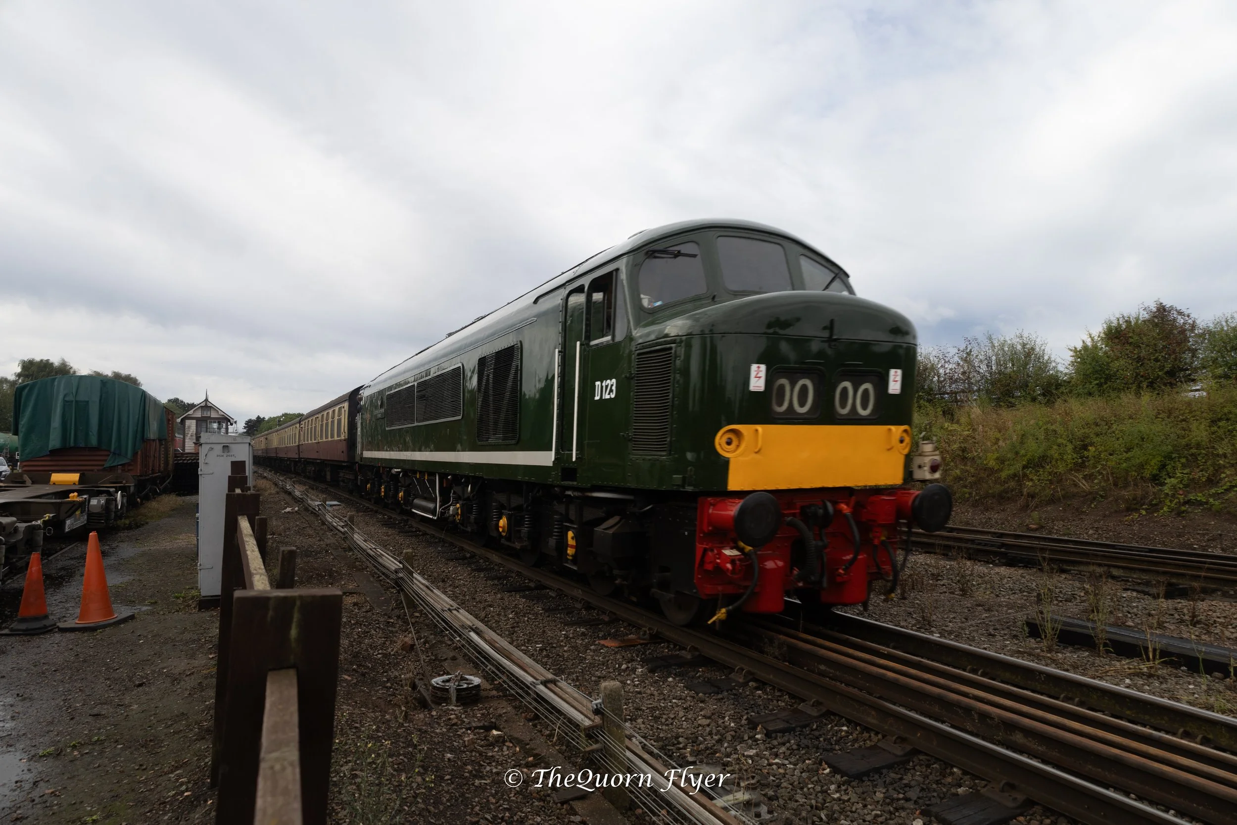 A green vintage train with a yellow front driving on railway tracks outdoors with trees and cloudy sky in the background.