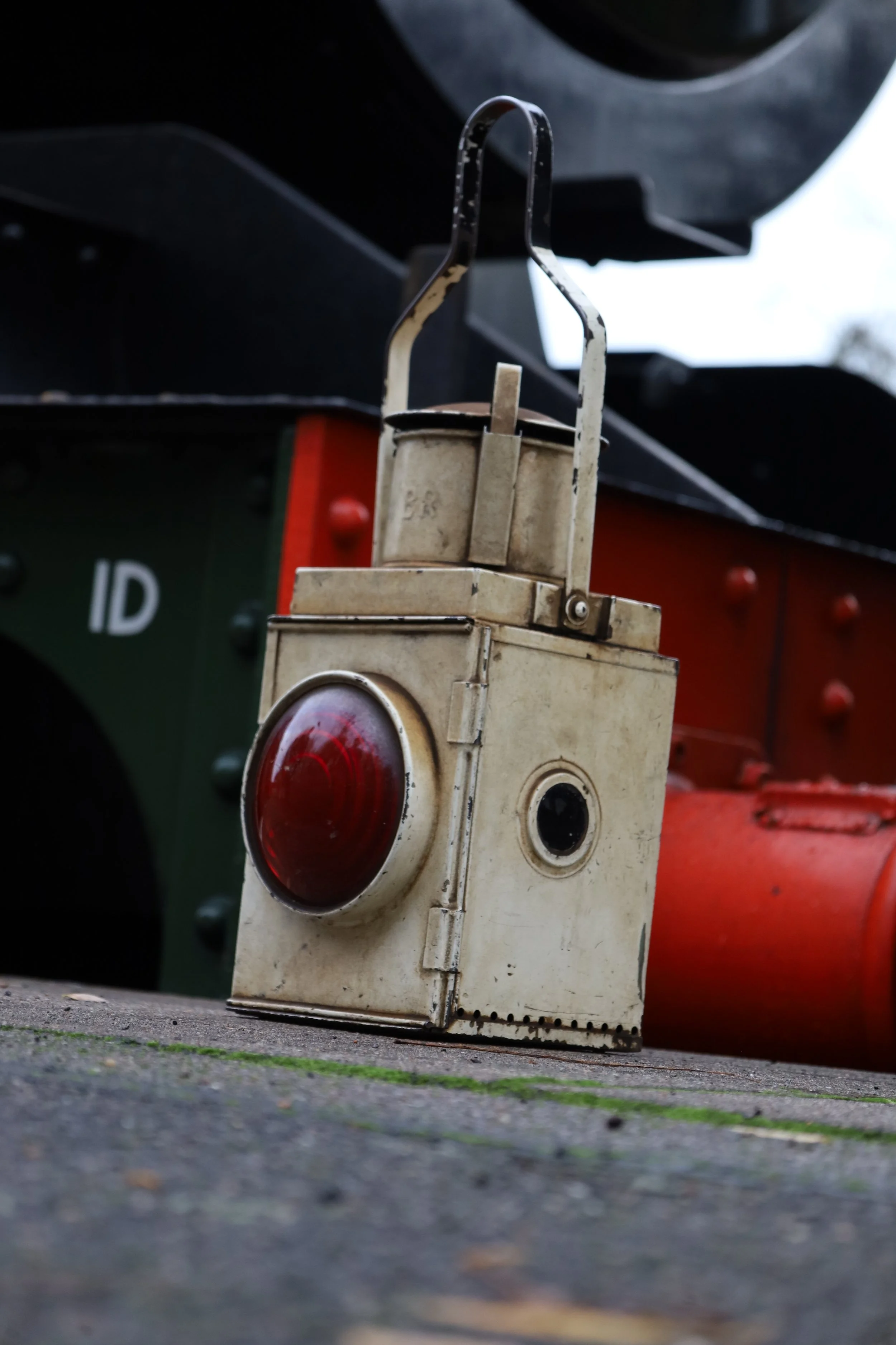 A close-up view of a vintage device with a large red circular light and metal components, positioned on a ground surface with a blurred background of a vehicle or machinery.