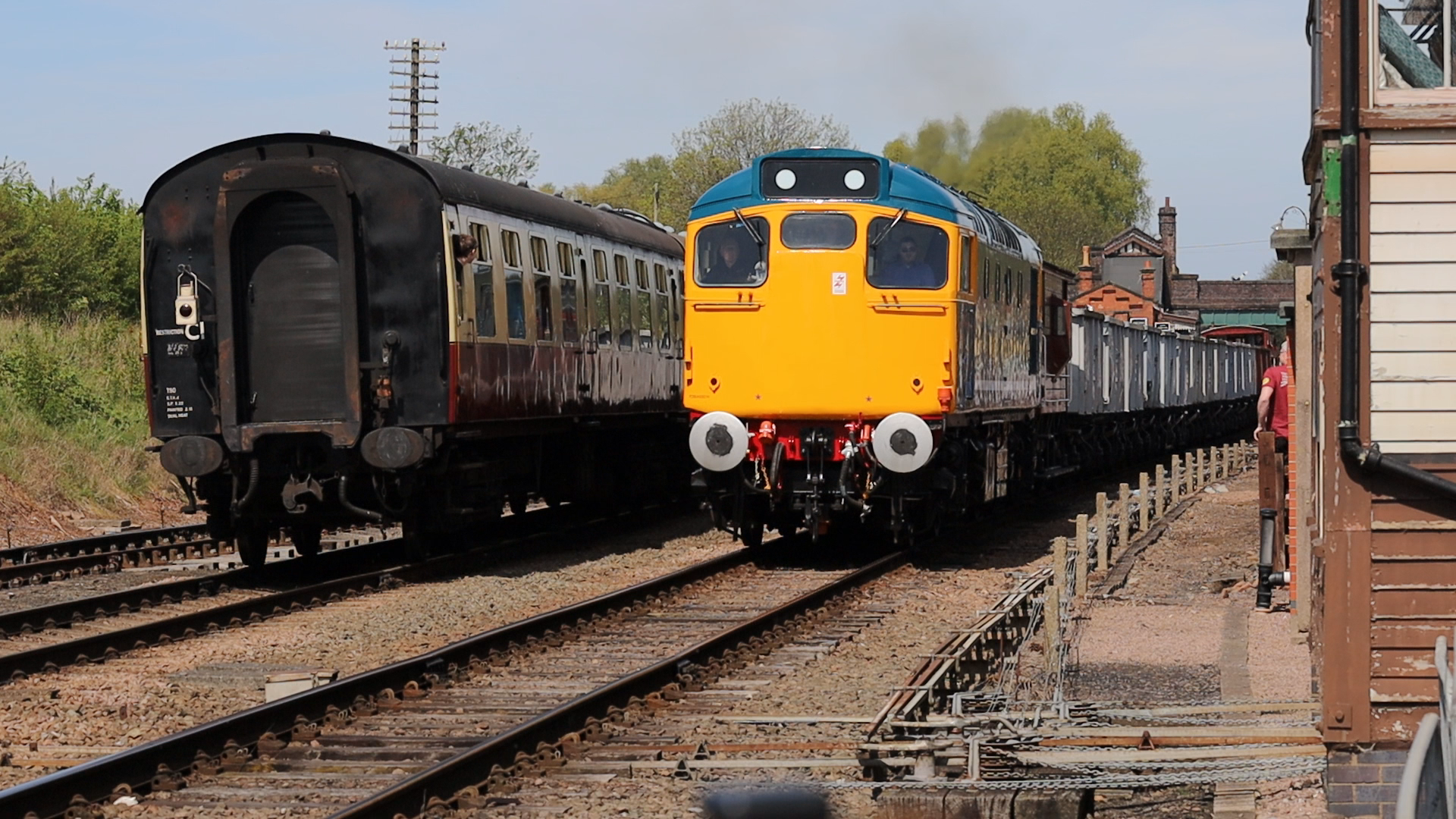 A vintage train with a yellow and blue locomotive pulling several brown passenger cars on railway tracks near a residential area with green trees and a clear sky.