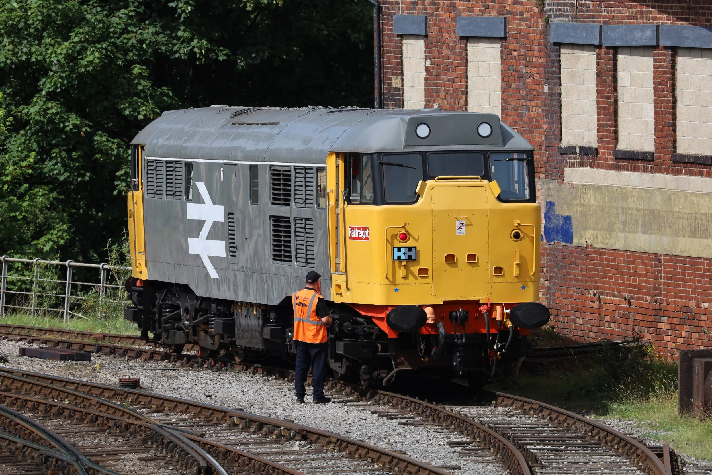 A train carriage under maintenance on railway tracks with two workers dressed in orange safety clothing inspecting it near a brick building.