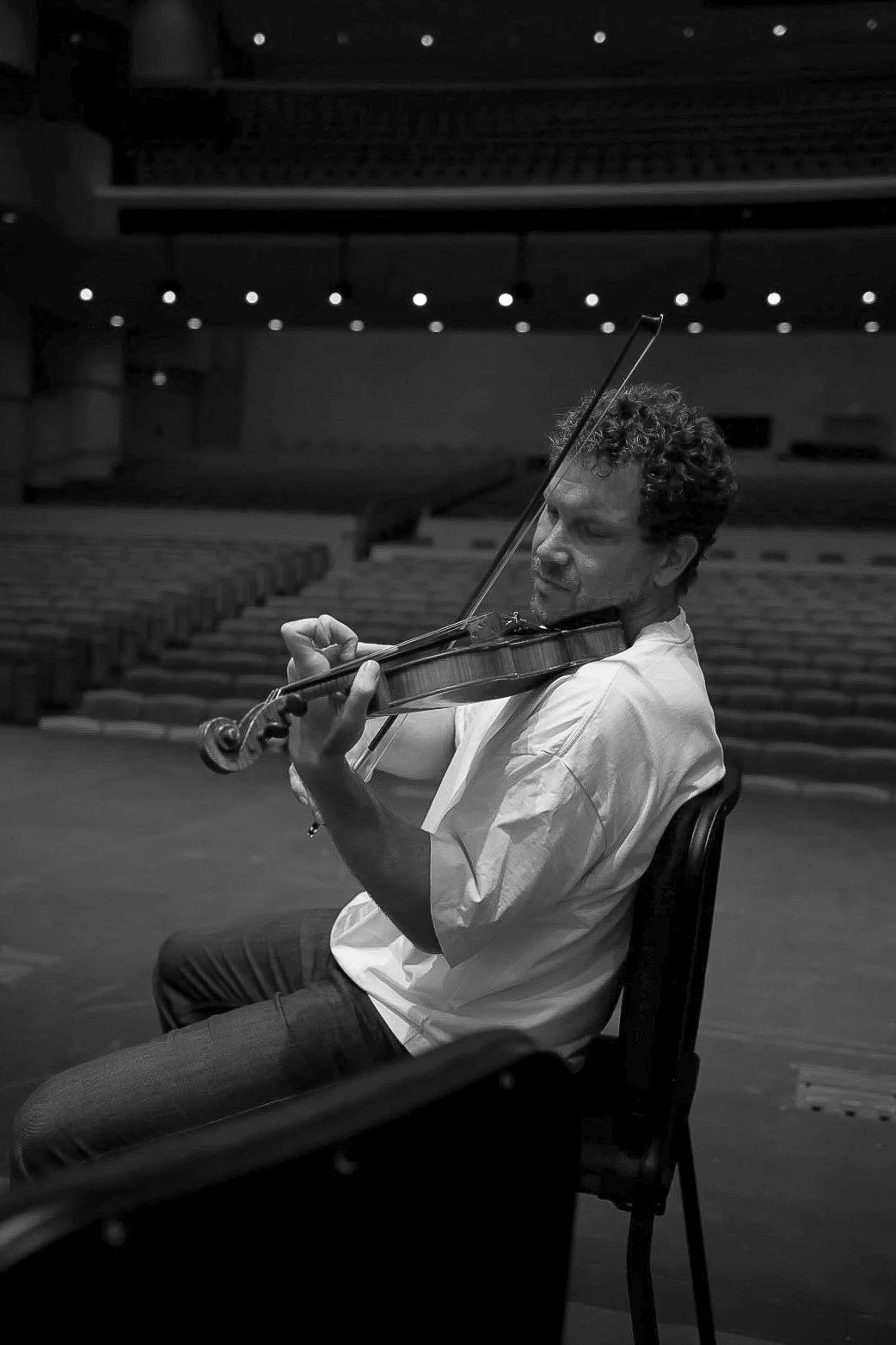 Black-and-white portrait of a seated violinist playing in an empty concert hall
