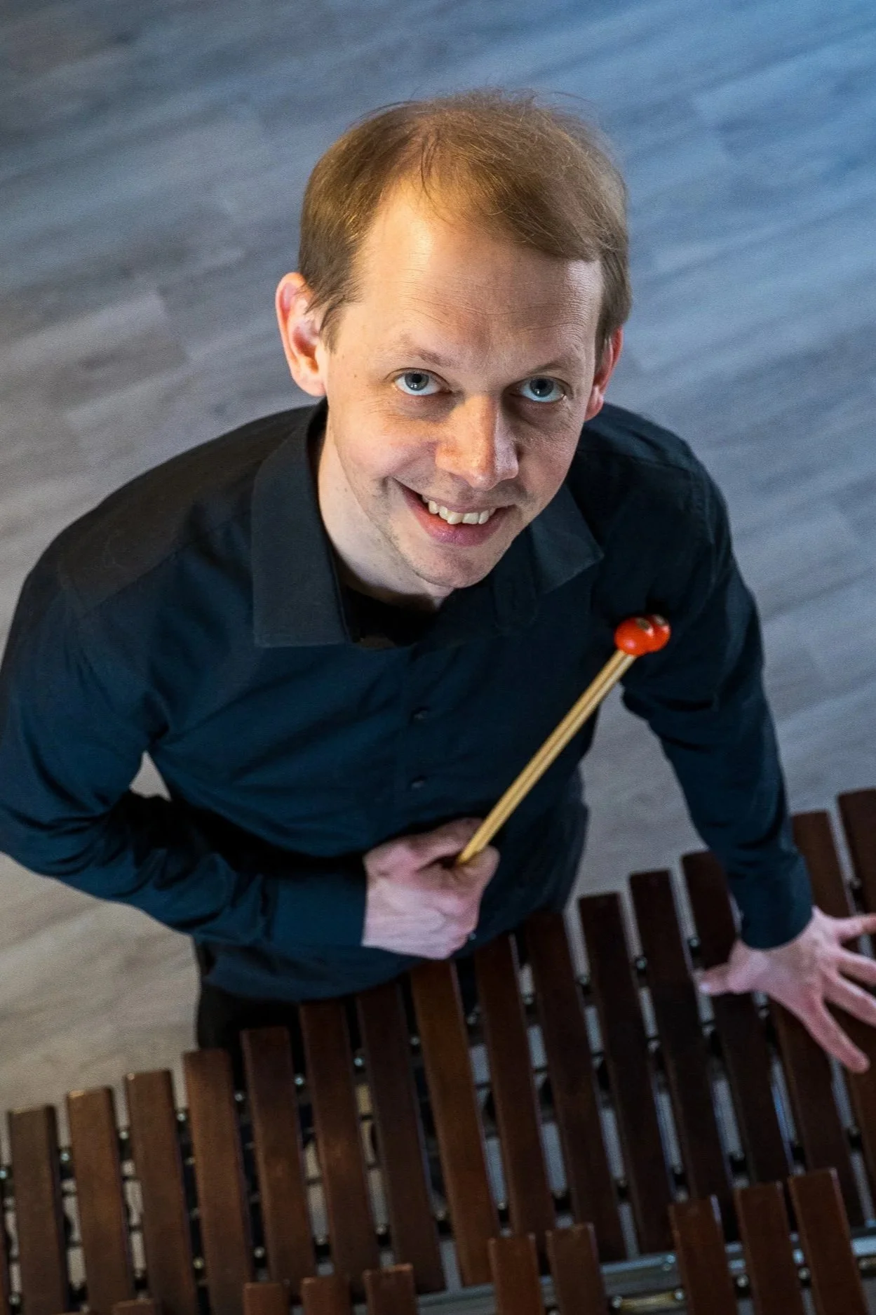 Smiling man in a black shirt holding mallets over a wooden xylophone