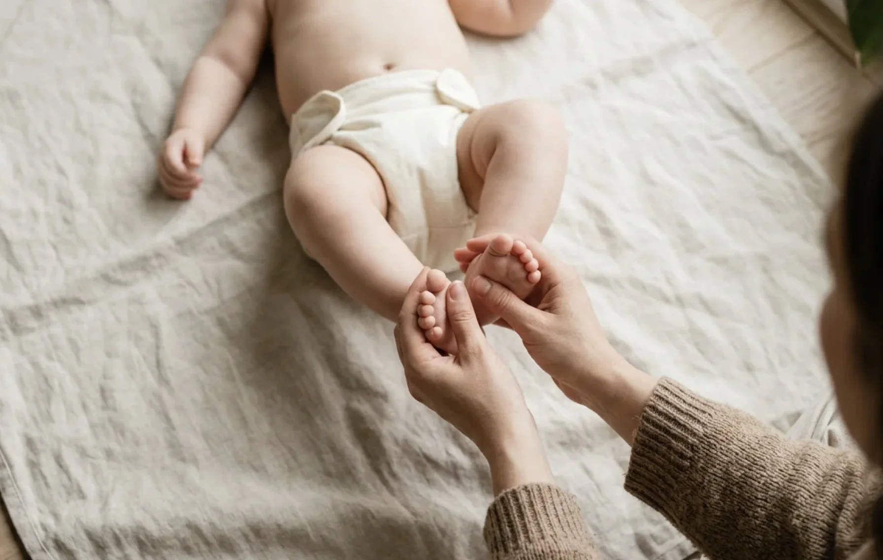 Näheaufnahme eines Babys in Windelhose, das auf einem Bett liegt, während eine erwachsene Person dessen Zehen hält.