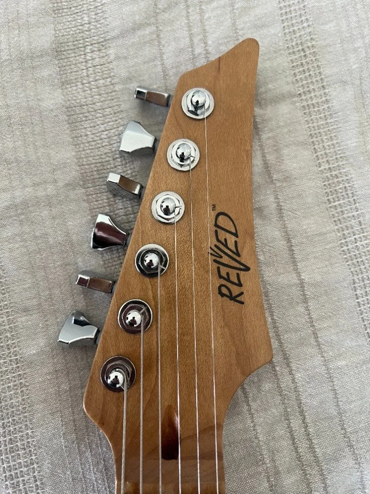 Close-up of a guitar headstock with tuning pegs and the brand name 'Revved' on a textured fabric background.