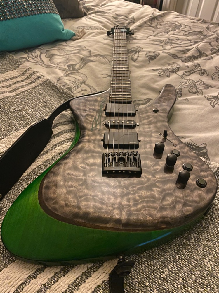 A green and black electric guitar with a quilted top, lying on a bed with patterned sheets and pillows.