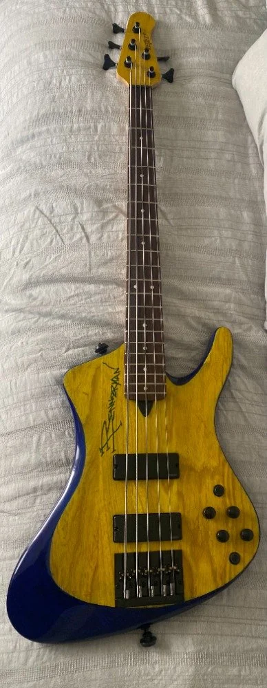 Yellow and blue electric bass guitar with a signed body, resting on a bed.