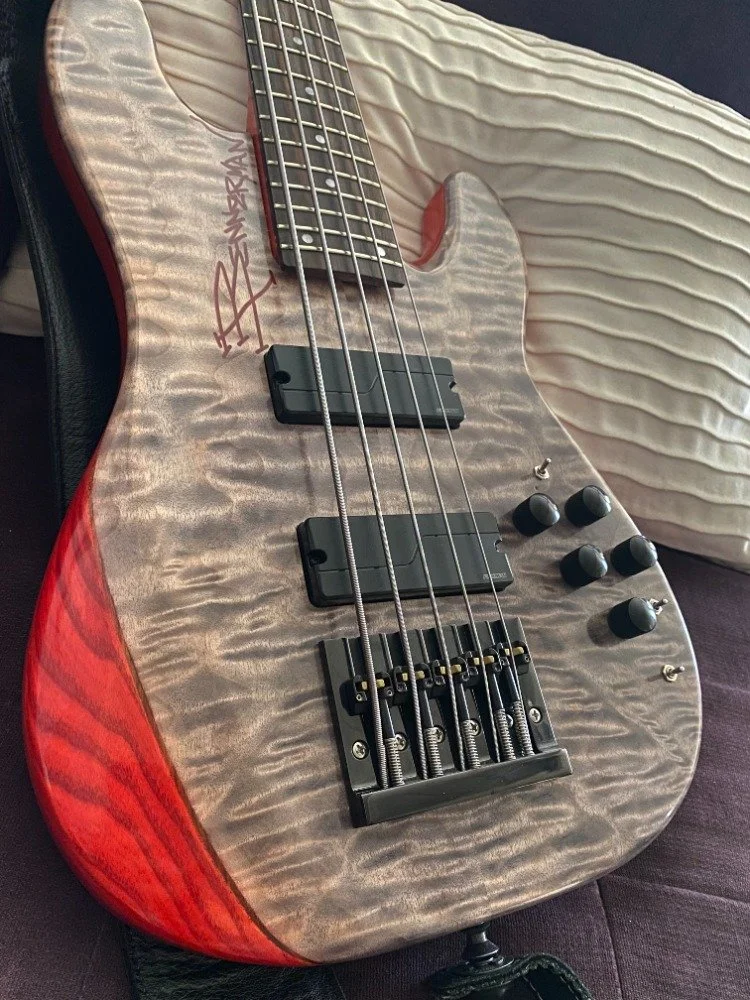 A close-up of a quilted electric bass guitar with a unique wood grain finish, black pickups, four control knobs, and a signed body resting on a textured surface.