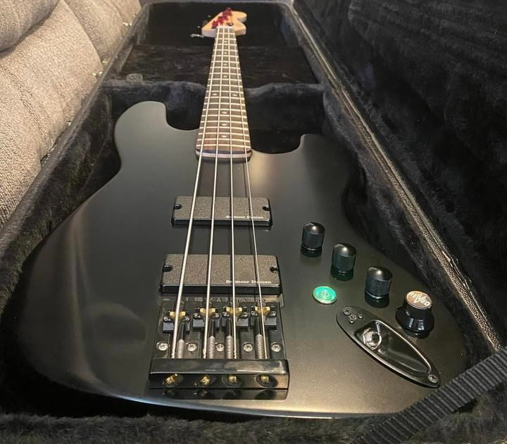 Black electric bass guitar inside a hardshell case, with control knobs and pickups visible, lying on a soft interior lining.