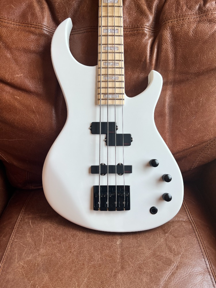 White electric bass guitar with a light wood neck, black pickups, and control knobs, resting on a brown leather couch.