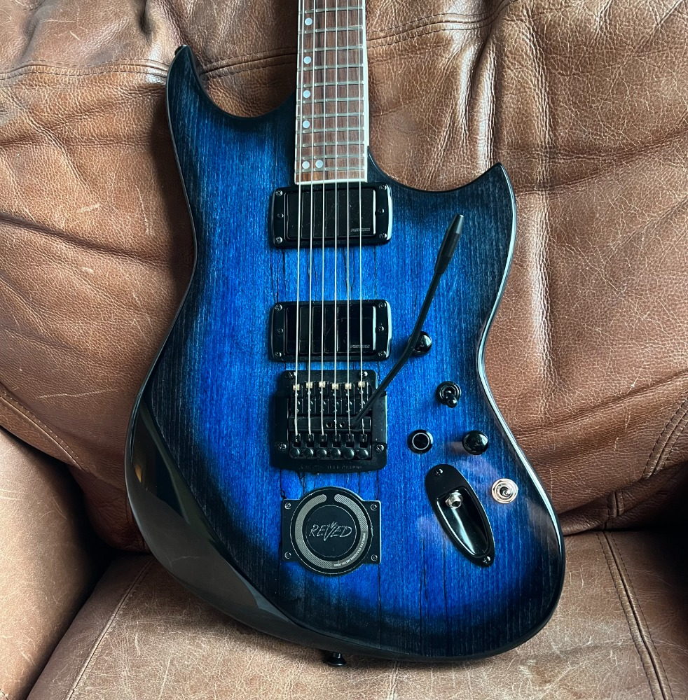 Blue electric guitar with MagSafe compatible charging receiver, resting on a brown leather sofa.