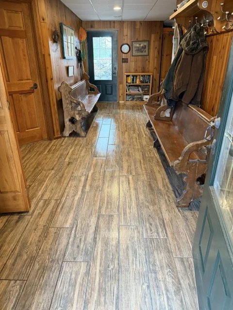 A cozy entryway with wood-paneled walls and hardwood flooring, featuring two wooden benches on either side, a small bookshelf, and a dark door with windows at the end.