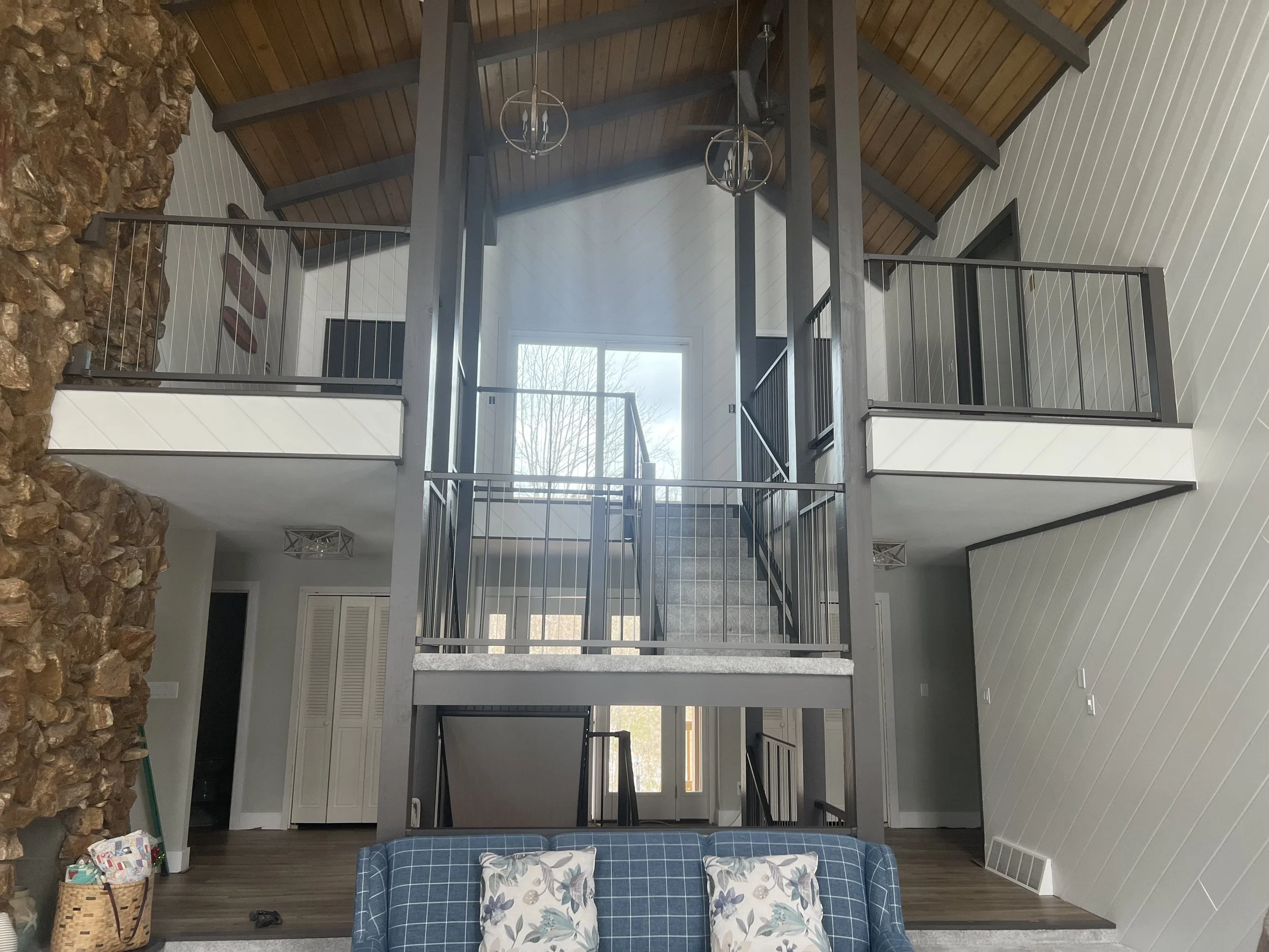 Interior view of a home showing a staircase with metal railings, a stone wall on the left, white paneled walls, and a high wood ceiling with hanging chandeliers. A sliding glass door lets in natural light, and part of a blue plaid couch with pillows is visible in the foreground.