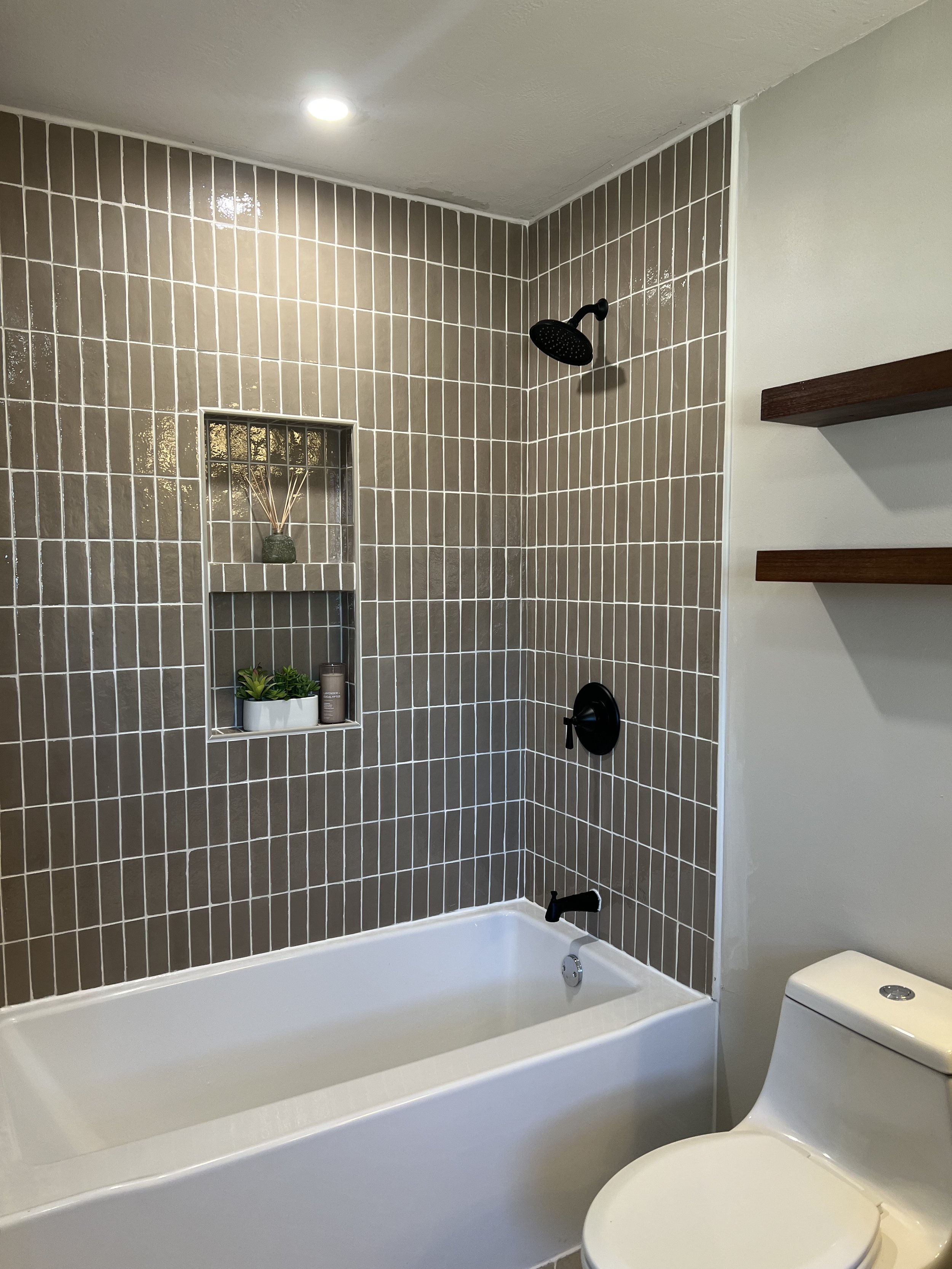 Bathroom shower with beige tile walls, black showerhead, and built-in shelf with potted plant and decorative items. White bathtub and a partial view of a white toilet.