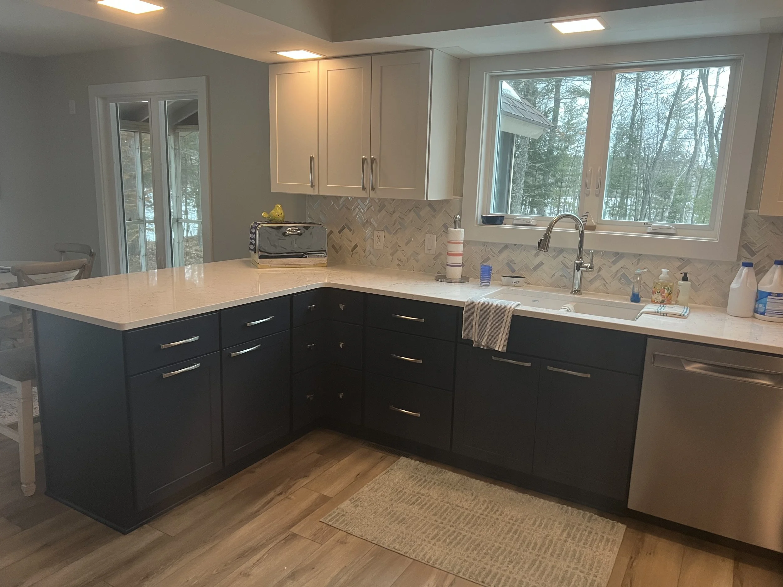 A kitchen interior with dark lower cabinets, light upper cabinets, a marble countertop, a large window showing trees outside, and kitchen utensils and soap bottles on the counter.