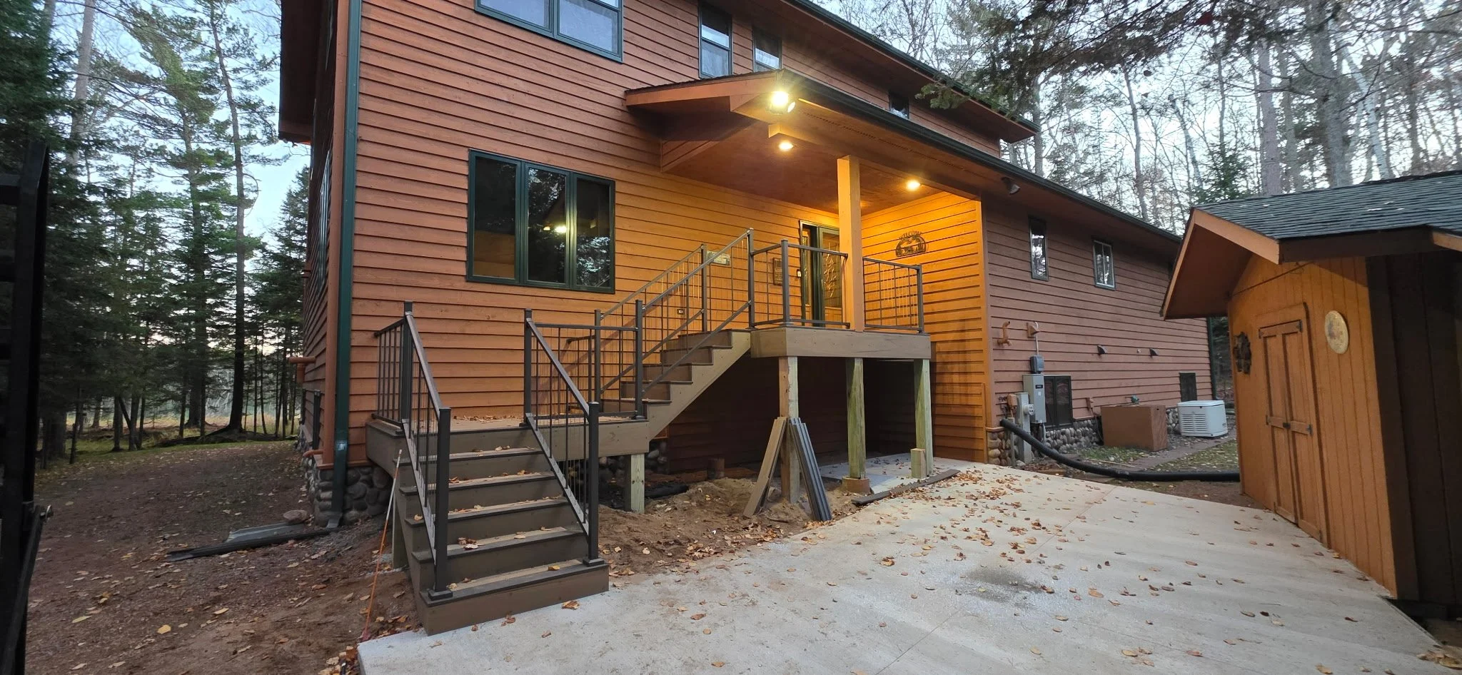 A two-story wooden house with a new staircase leading to a small porch. The house has warm lighting, large windows, and is surrounded by trees in a wooded area.