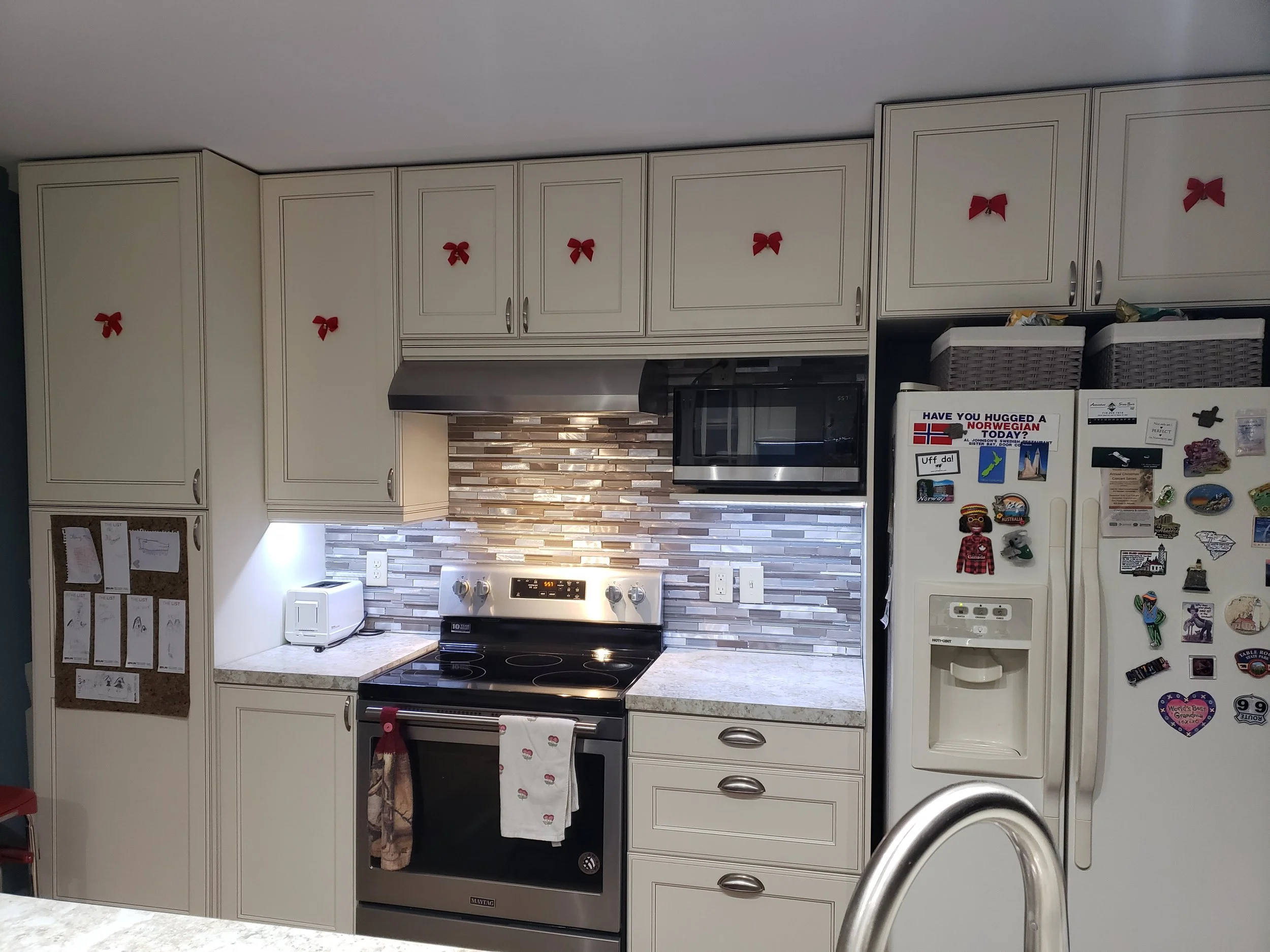 Kitchen with white cabinets decorated with red bows, a stove, microwave, refrigerator with various magnets, and a tiled backsplash.