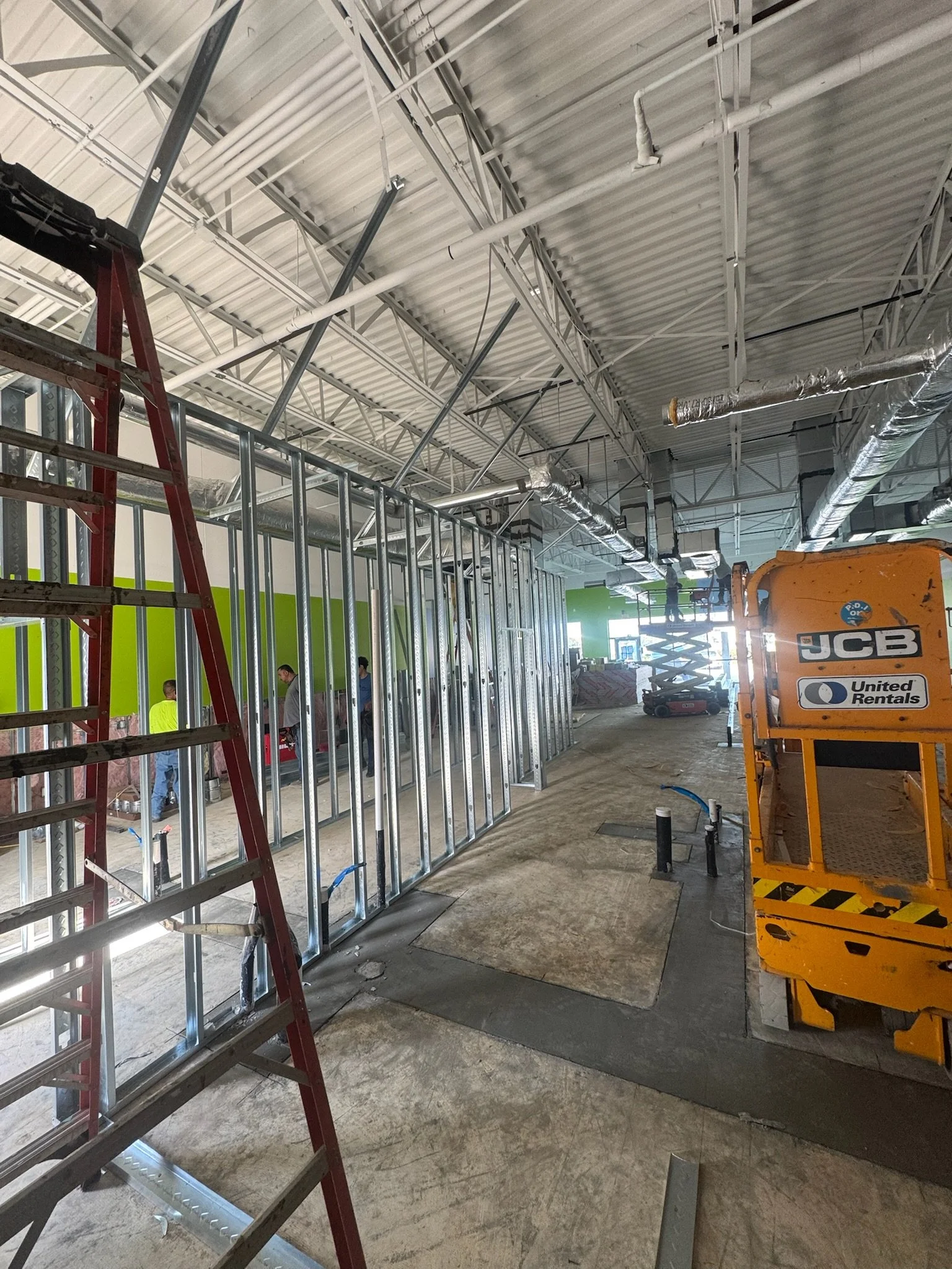 Construction site indoors with unfinished walls and ceiling, metal framing, scaffolding, utility pipes, and construction equipment.