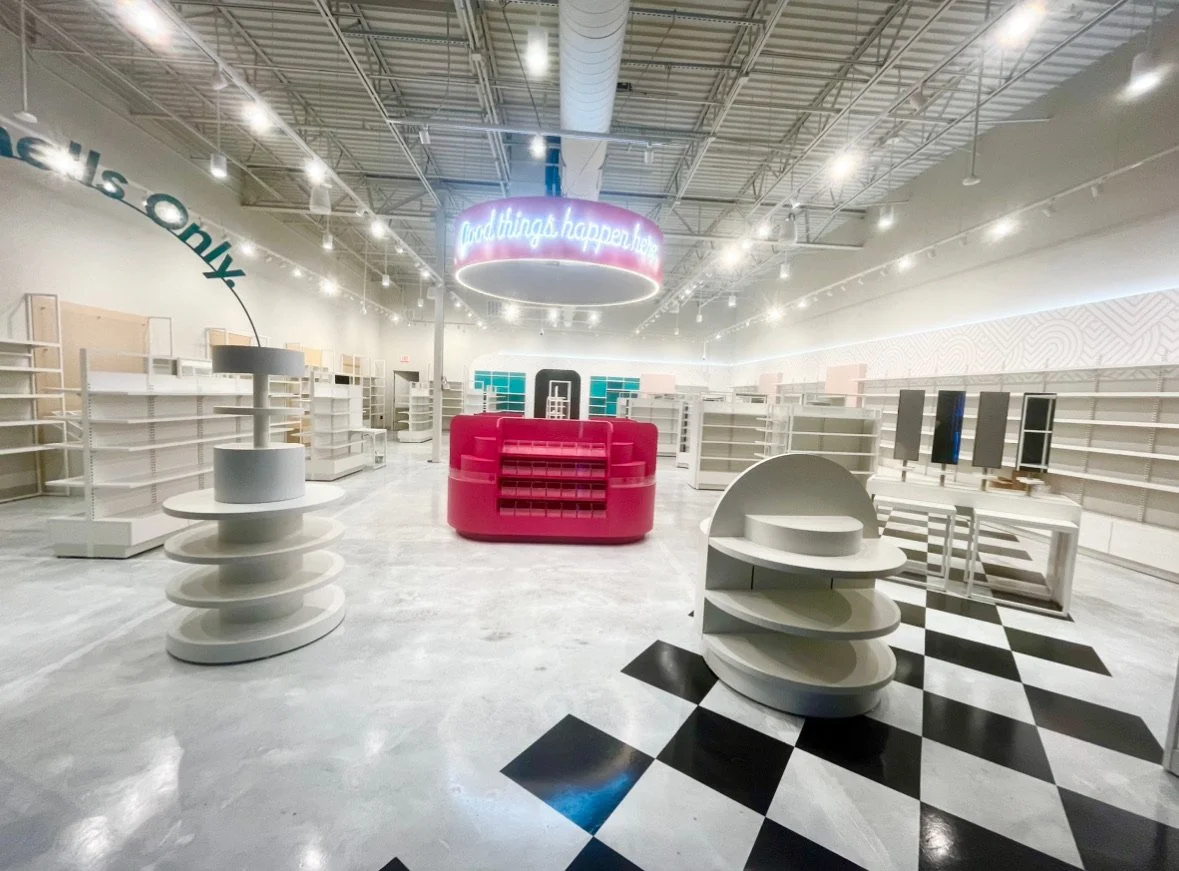 Empty retail store with white shelves, a central pink display, and a hanging circular sign that reads 'Good things happen here,' with a black and white checkered floor section.