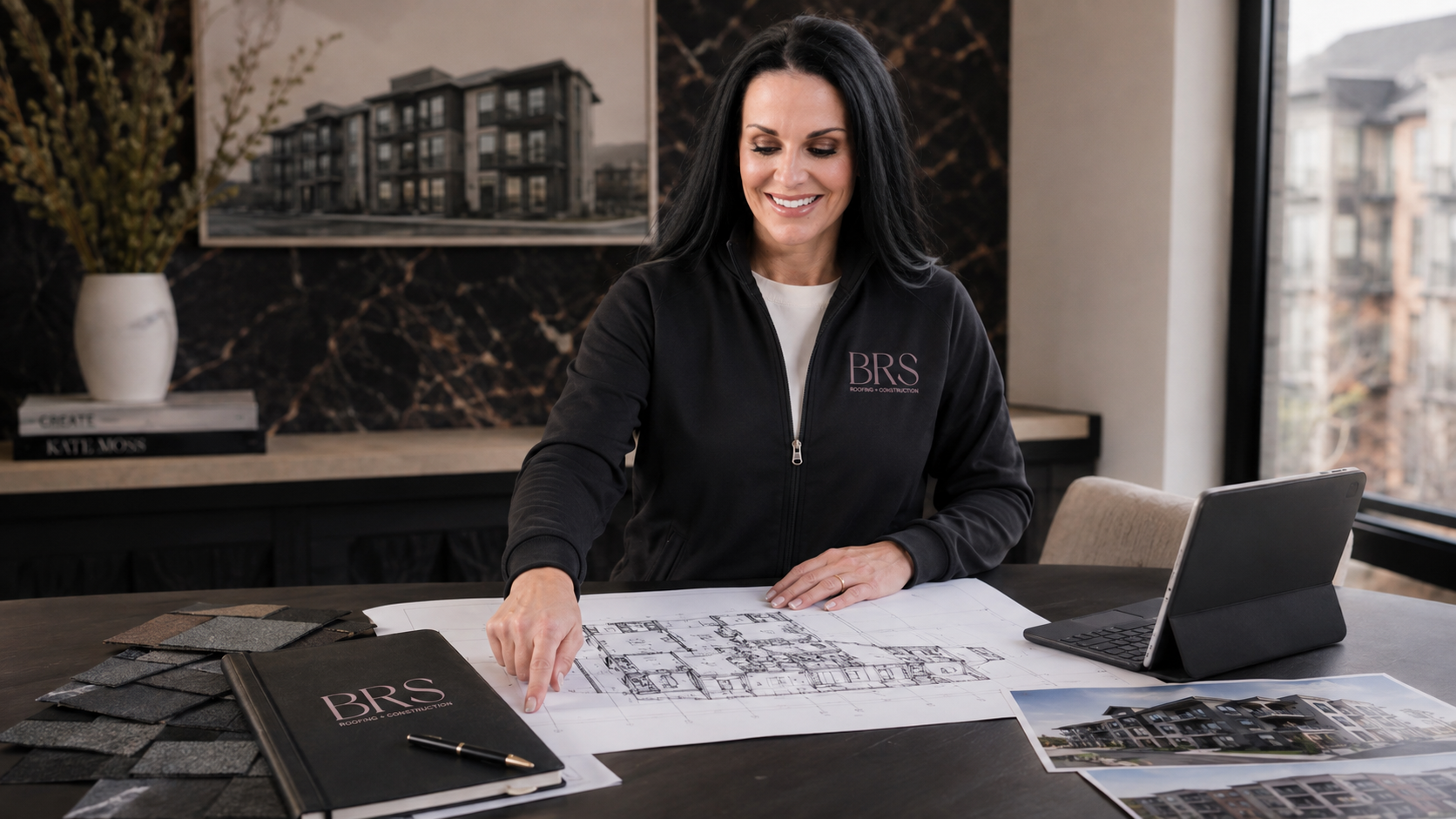 A smiling woman with black hair working on architectural plans at a table with design materials, a laptop, and a photo of a building, in a modern office with large windows and a city view.