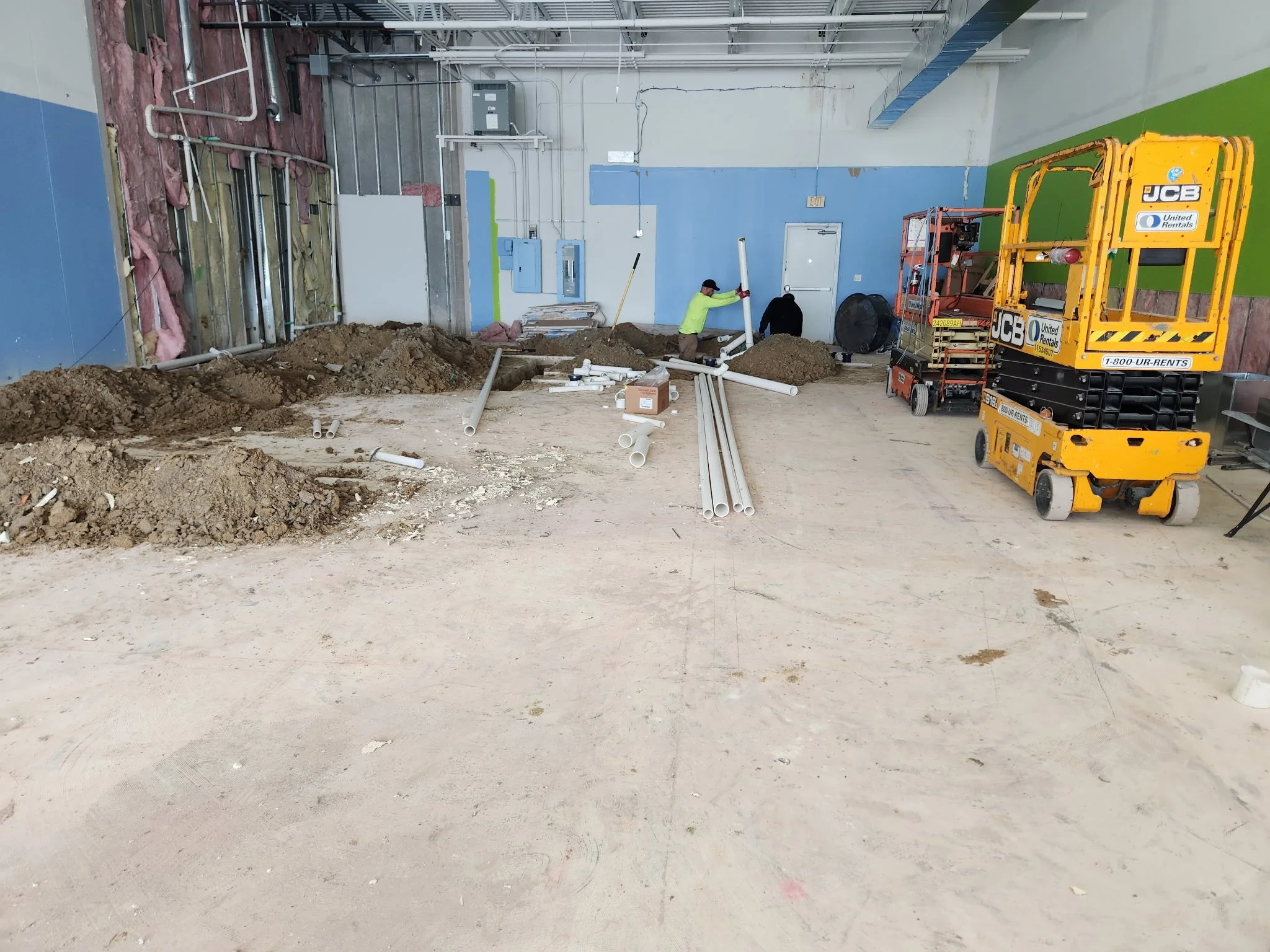 Construction workers installing pipes in an indoor construction site with piles of dirt, construction equipment, and partially finished walls.