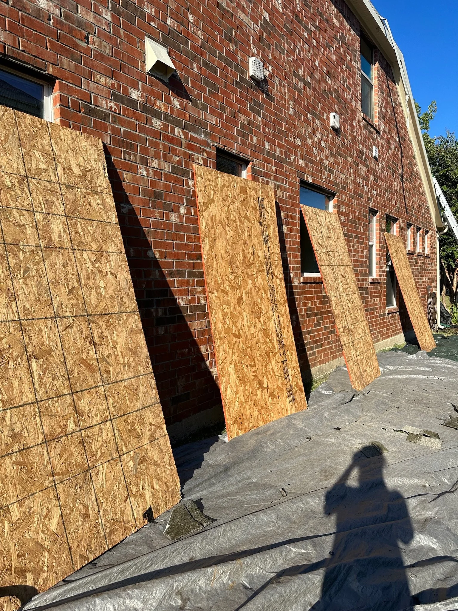 Construction site with a brick building and plywood sheets leaning against it, with a protective ground cover and sunlight casting shadows.