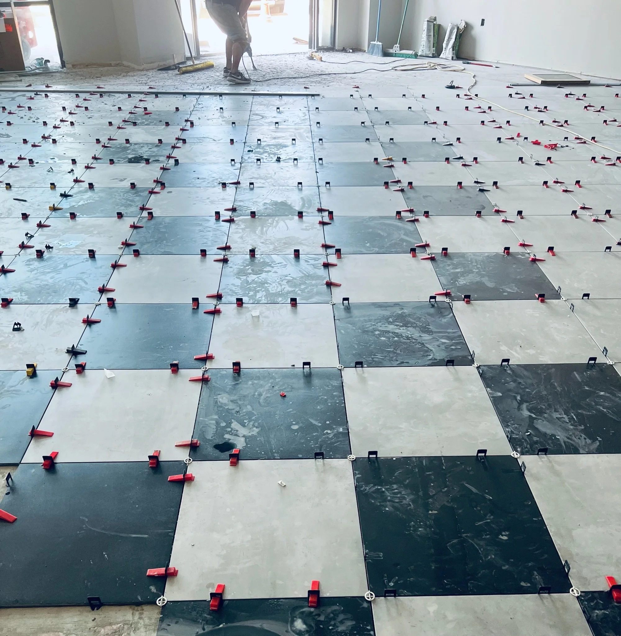 A person installing ceramic tiles on a floor in a room under construction or renovation, with tools and materials visible in the background.