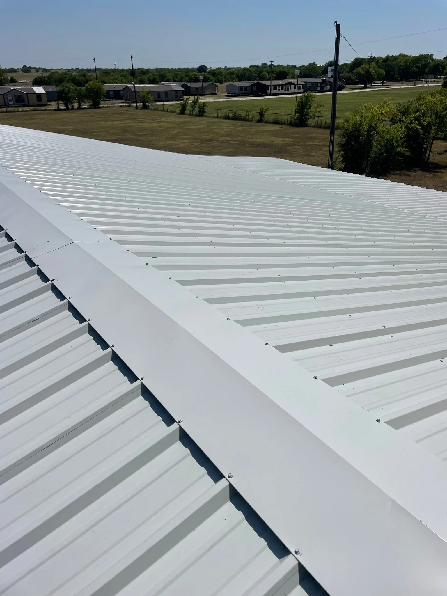 Close-up of a white metal roof with panels and screws, with a background of a grassy field, trees, utility poles, and a clear blue sky.